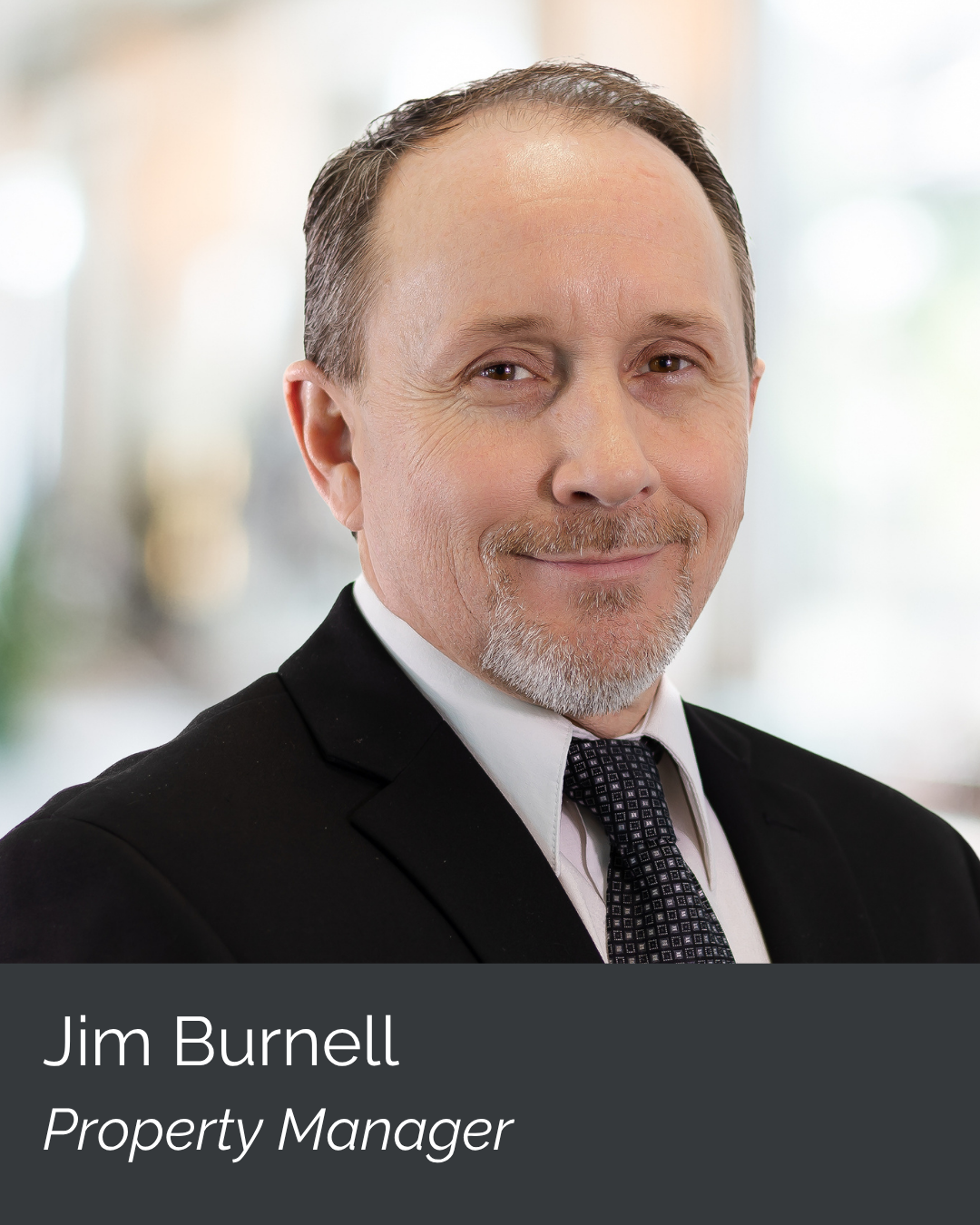 A middle-aged man with short gray hair and a beard, dressed in a black suit, white shirt, and patterned tie, smiling at the camera. Background is blurred. Text overlay reads: "Jim Burnell, Property Manager."