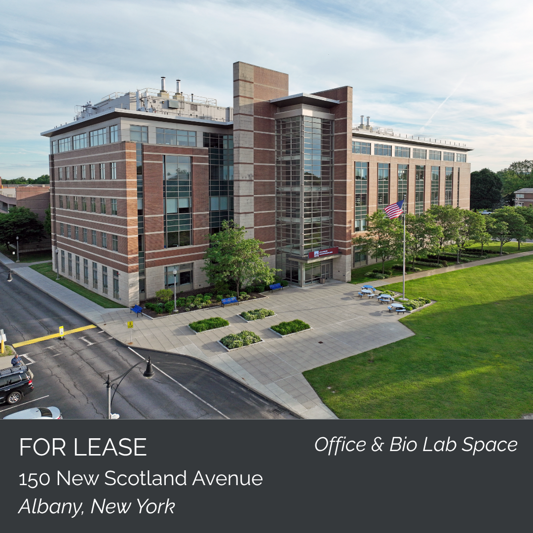 A modern office building with brick and glass architecture, surrounded by green grass, trees, a sidewalk, and a flagpole with an American flag, located at 150 New Scotland Avenue in Albany, New York, available for lease as office and bio lab space.