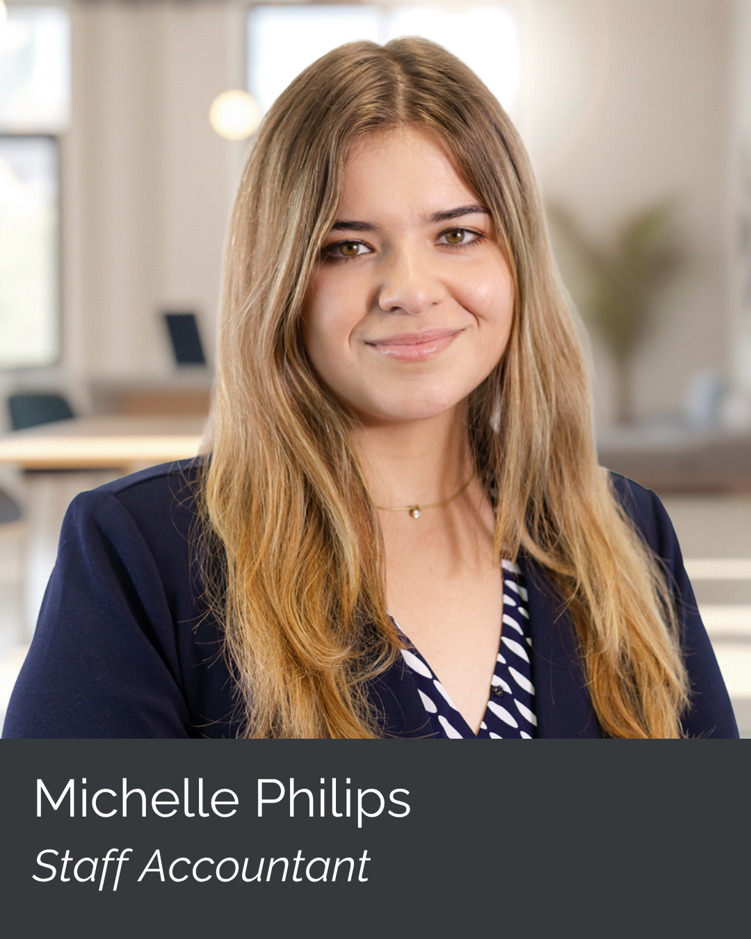 Gender image of a young woman with long, wavy brown hair, smiling, wearing a navy blazer and a patterned blouse, in a well-lit office environment.