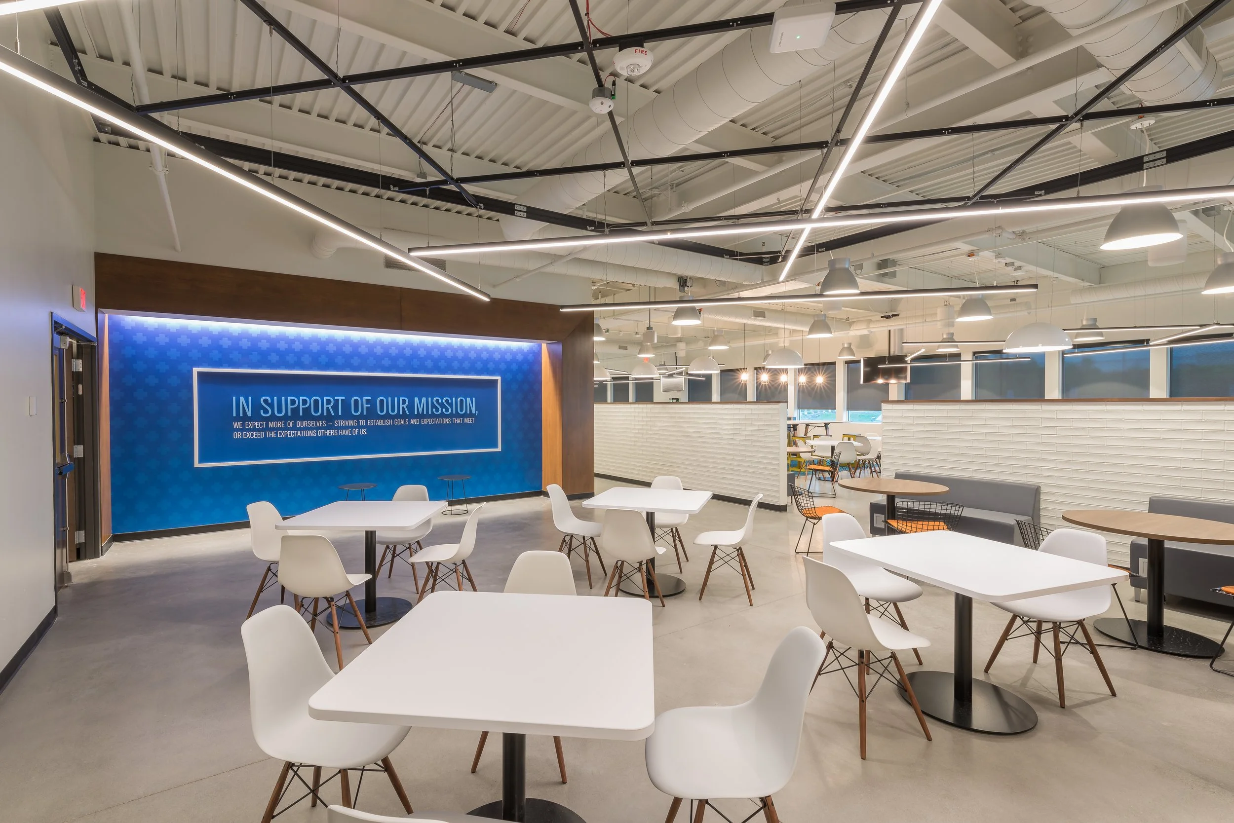 Modern open-concept coworking space with white tables, white chairs, and a lounge area with armchairs. The ceiling has exposed ducts and black and white industrial lighting fixtures. A blue wall with a sign reads 'In Support of Our Mission'.