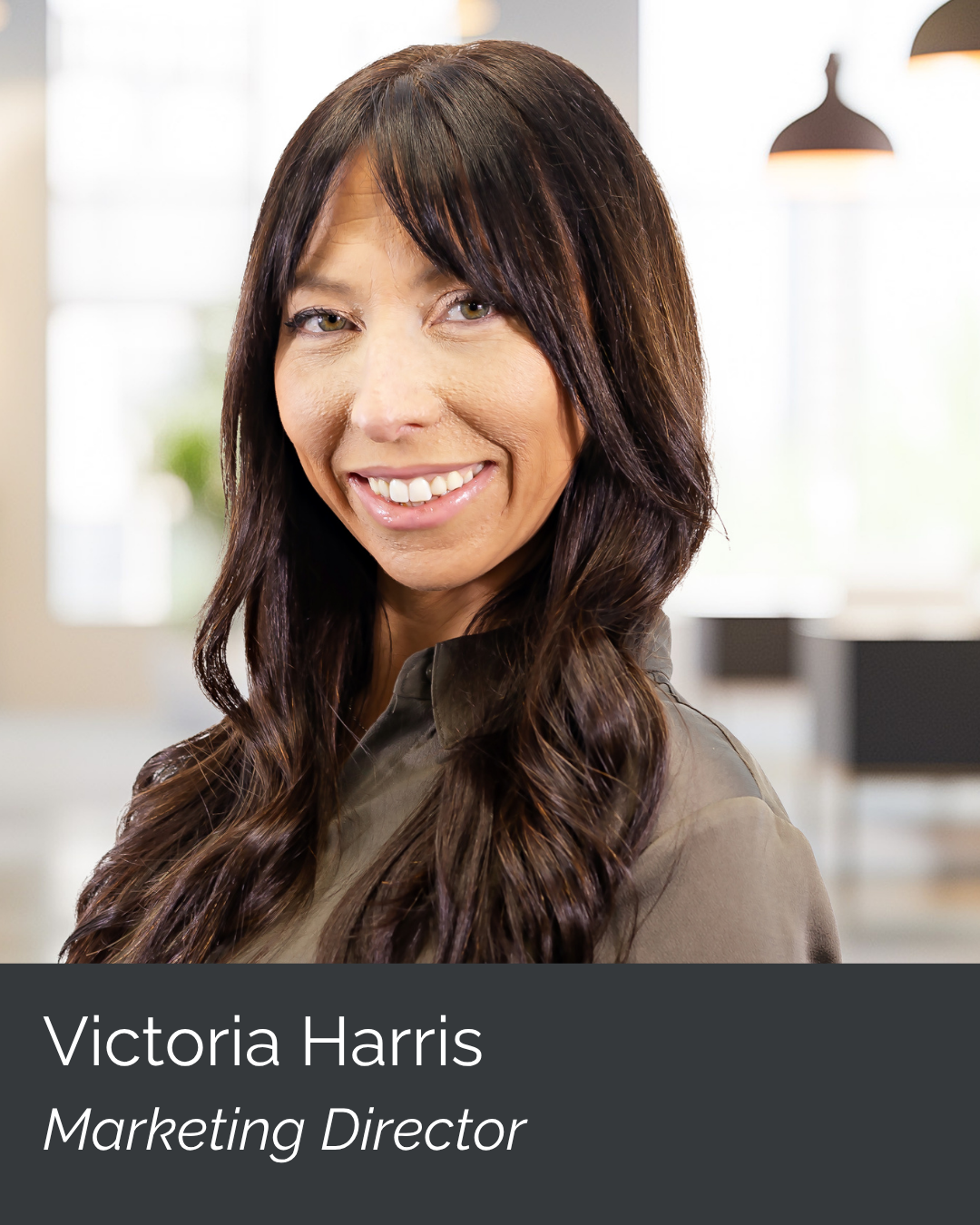 A woman with long dark brown hair and green eyes smiling in an office environment. She is wearing a gray blouse and is the marketing director, Victoria Harris.