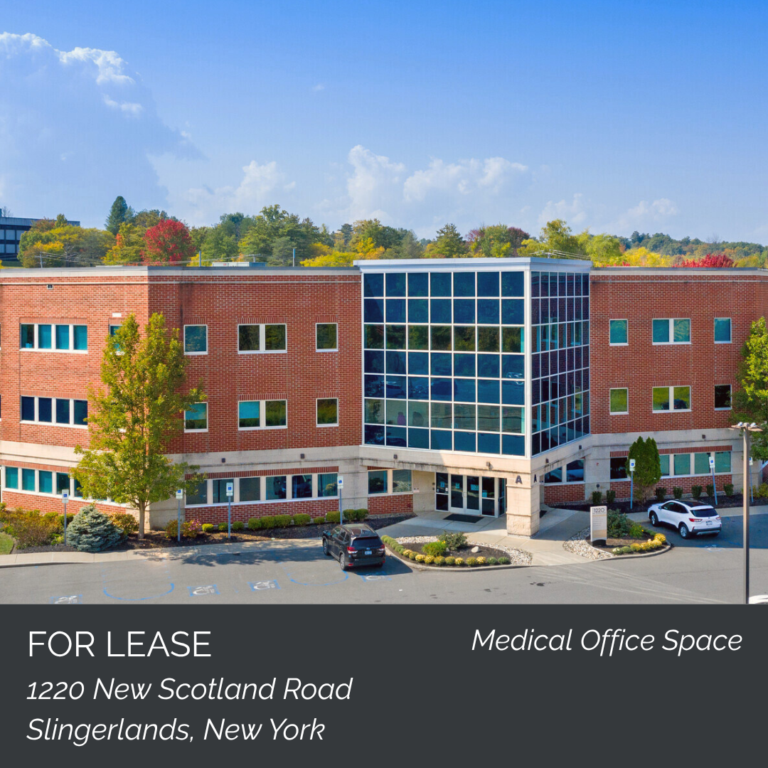 Exterior view of a modern medical office building with a brick facade and a glass entrance, parking lot in front, surrounded by trees and clear blue sky.