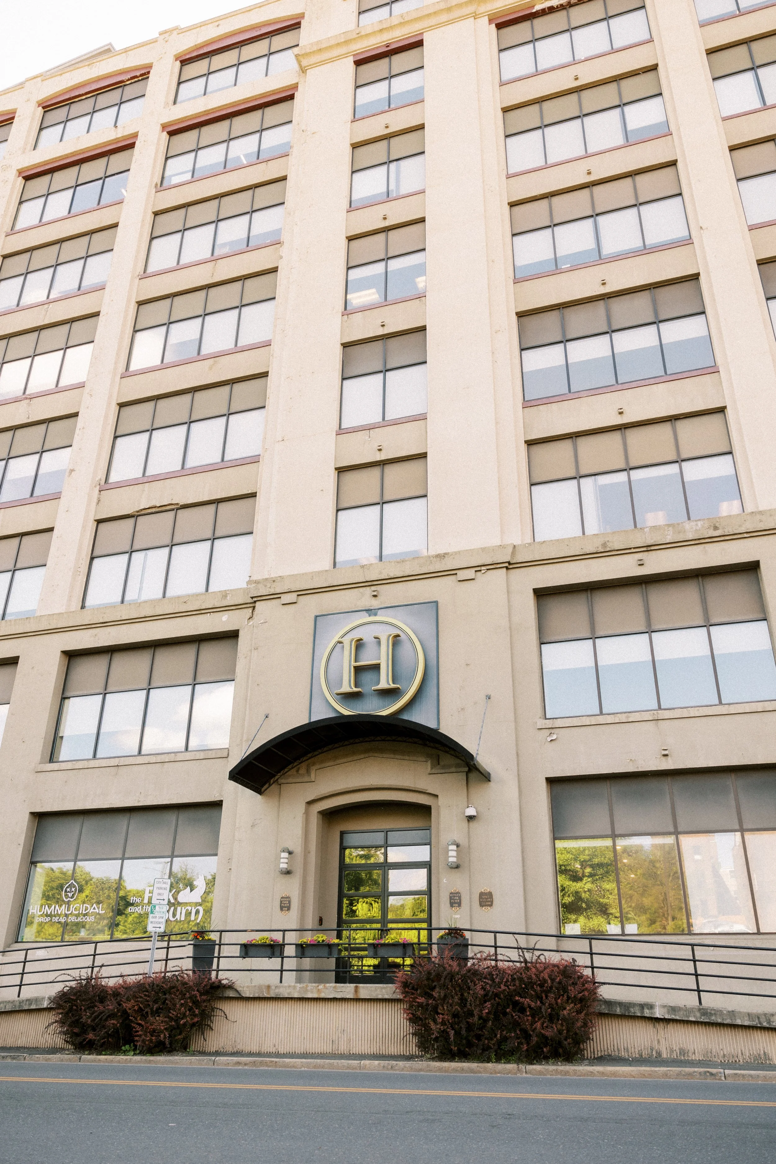 A multi-story beige building with large glass windows, a sign with the letter 'H' above the entrance, and a glass door. There are small plaques near the door, and a railing with potted plants in front. Bushes with reddish leaves are at the bottom of the building. The street is visible at the bottom of the image.