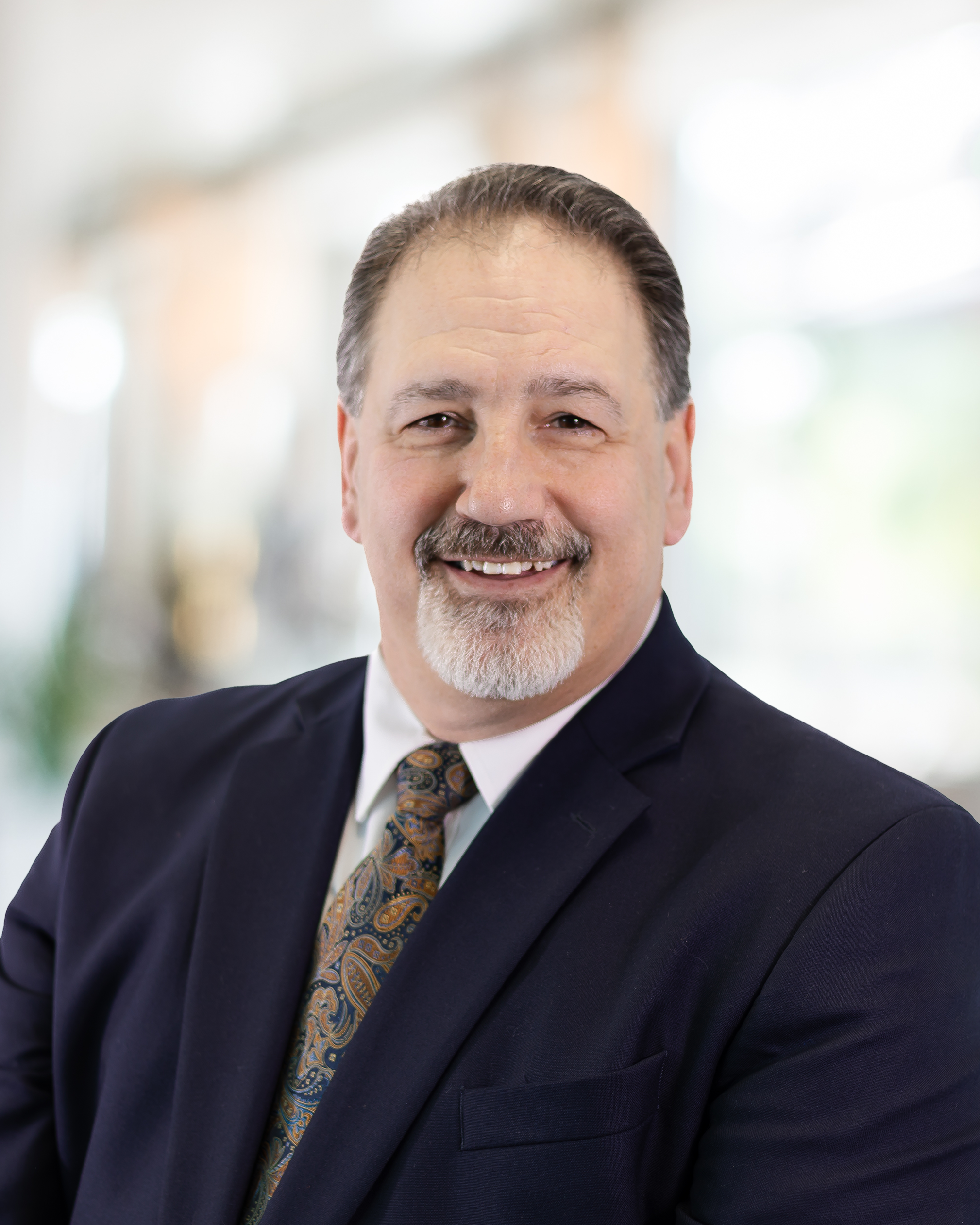 Close-up of a middle-aged man with dark hair, gray beard, and mustache, smiling and wearing a dark suit, white shirt, and patterned tie, with a blurred office background.