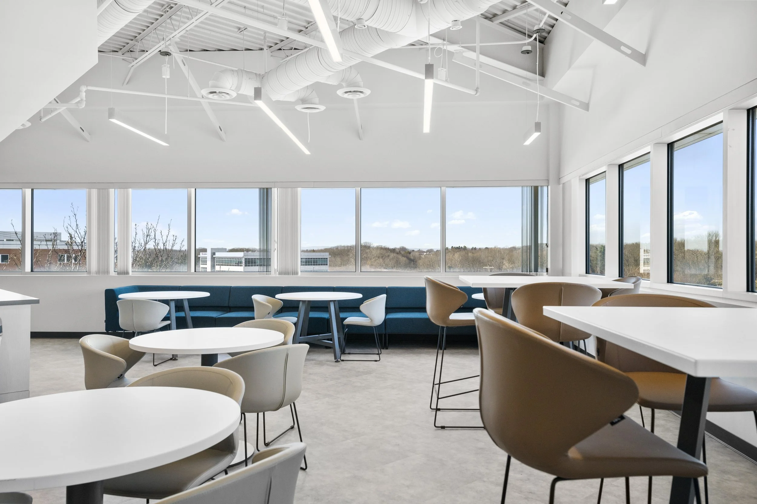 Modern interior of a bright office or lounge area with large windows, white tables, beige and white chairs, and a blue sofa. Ceiling features exposed ducts and linear lighting.
