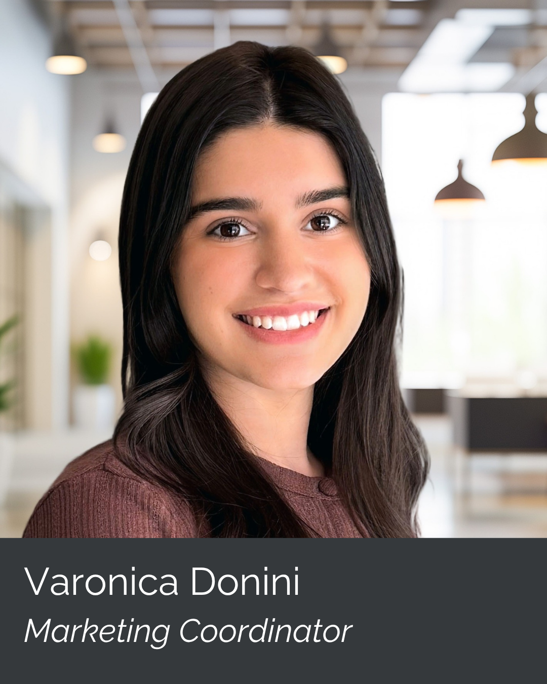 A young woman with long dark hair smiling in a bright modern office, wearing a brown top. Text overlay: Varonica Donini, Marketing Coordinator.