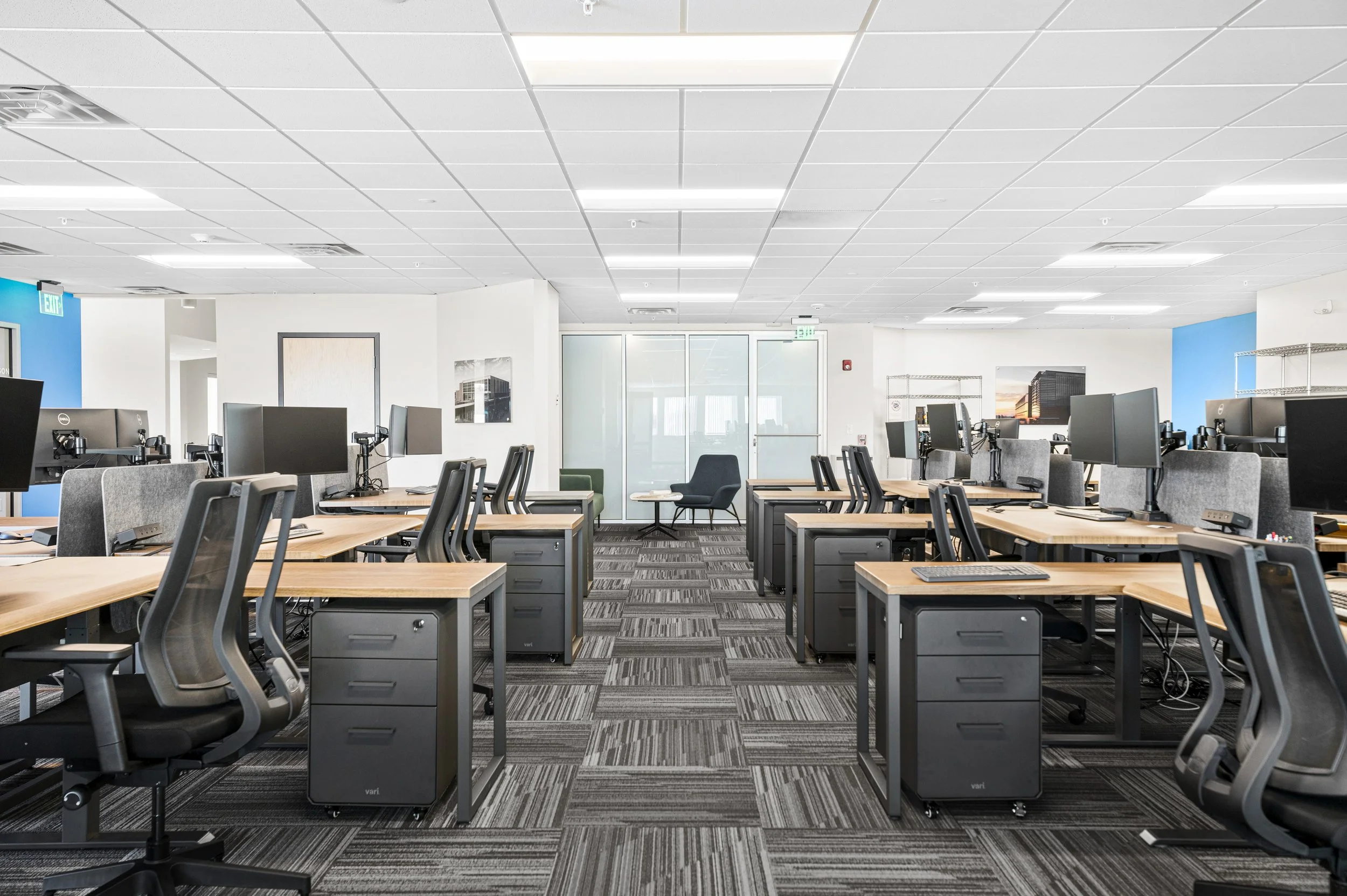 An empty modern office space with rows of desks, computers, and chairs in a well-lit room with a glass door at the back and blue accents on the walls.