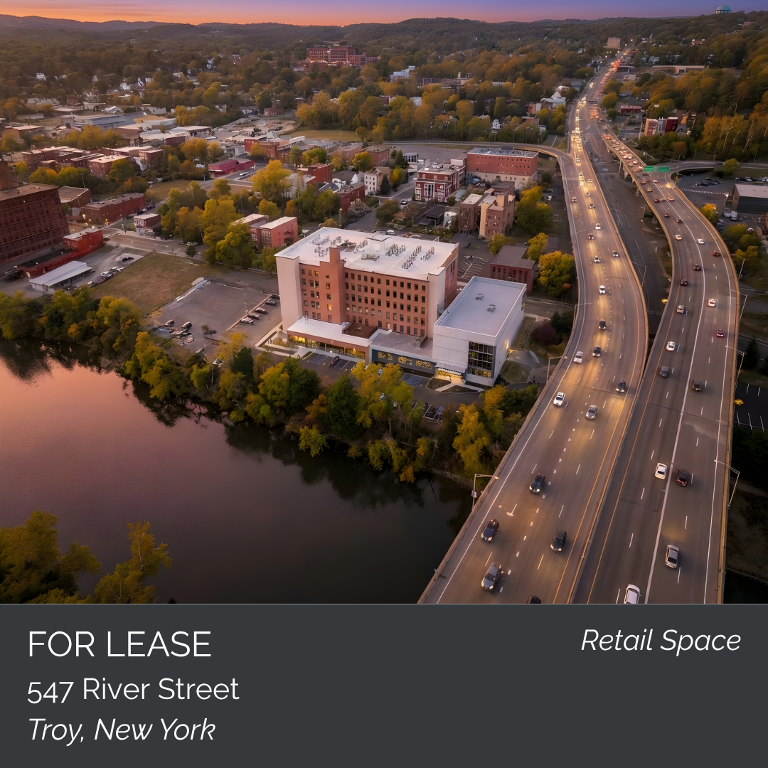An aerial view of Troy, New York, showing a large white commercial building near a body of water, with a highway nearby and a mix of trees and smaller buildings in the background during sunset.