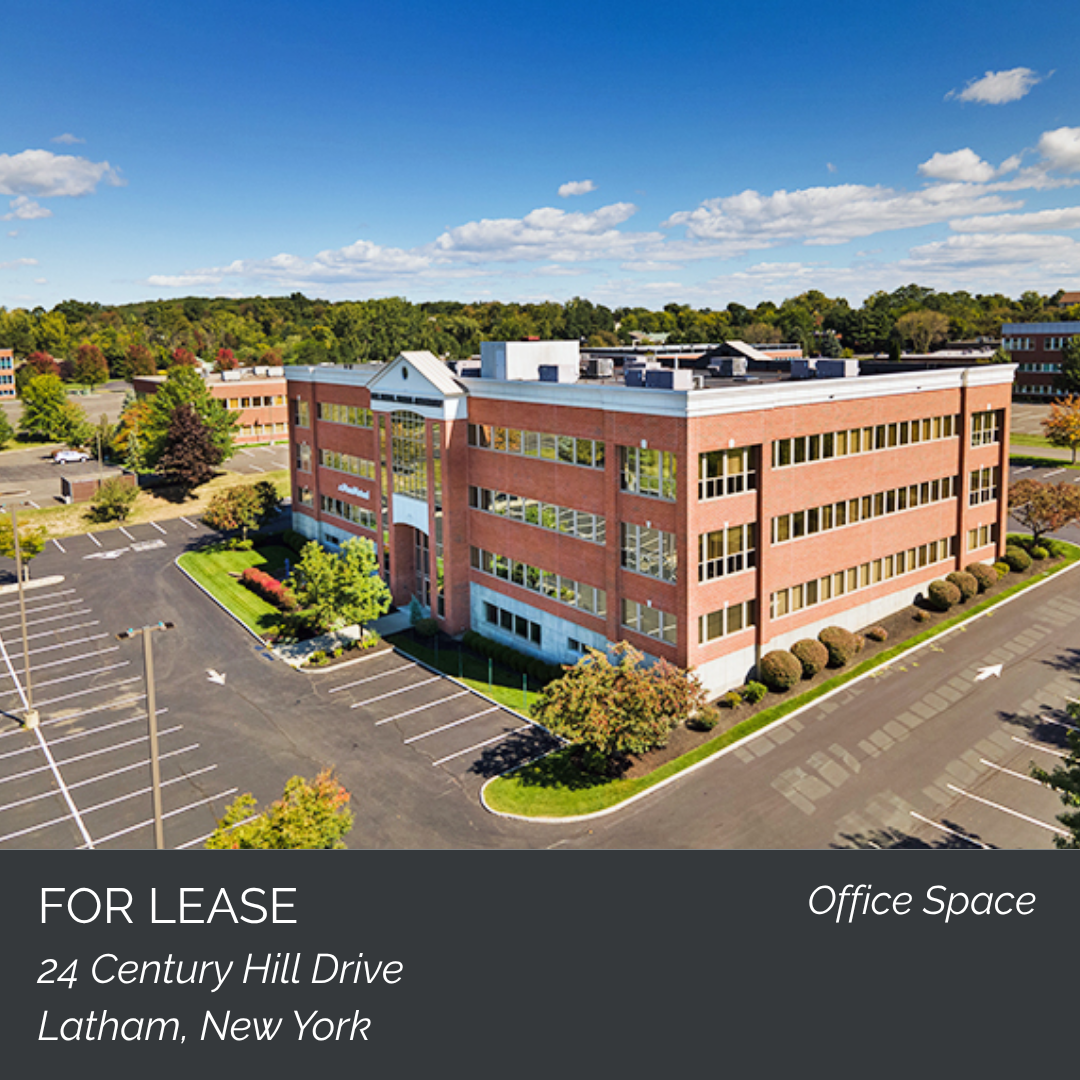 Aerial view of a three-story office building with a parking lot, surrounded by trees, under a blue sky with clouds. The building is located at 24 Century Hill Drive, Latham, New York, and is available for lease.