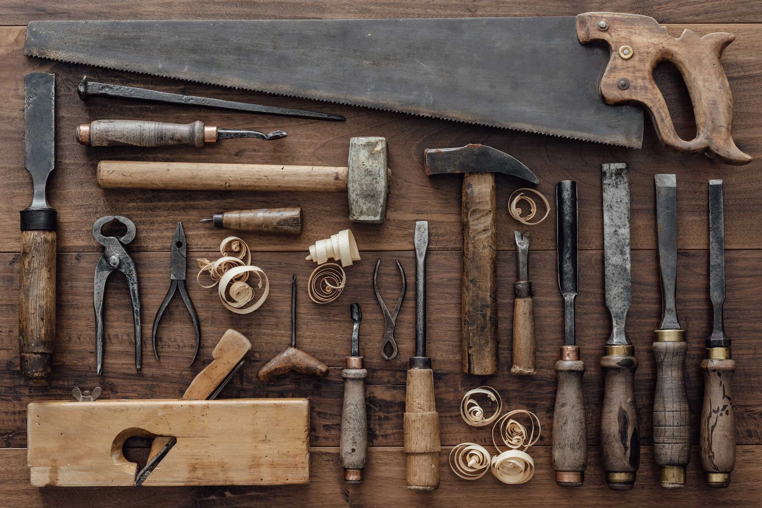 A collection of vintage woodworking tools, including saws, hammers, chisels, and carving tools, neatly arranged on a wooden surface.