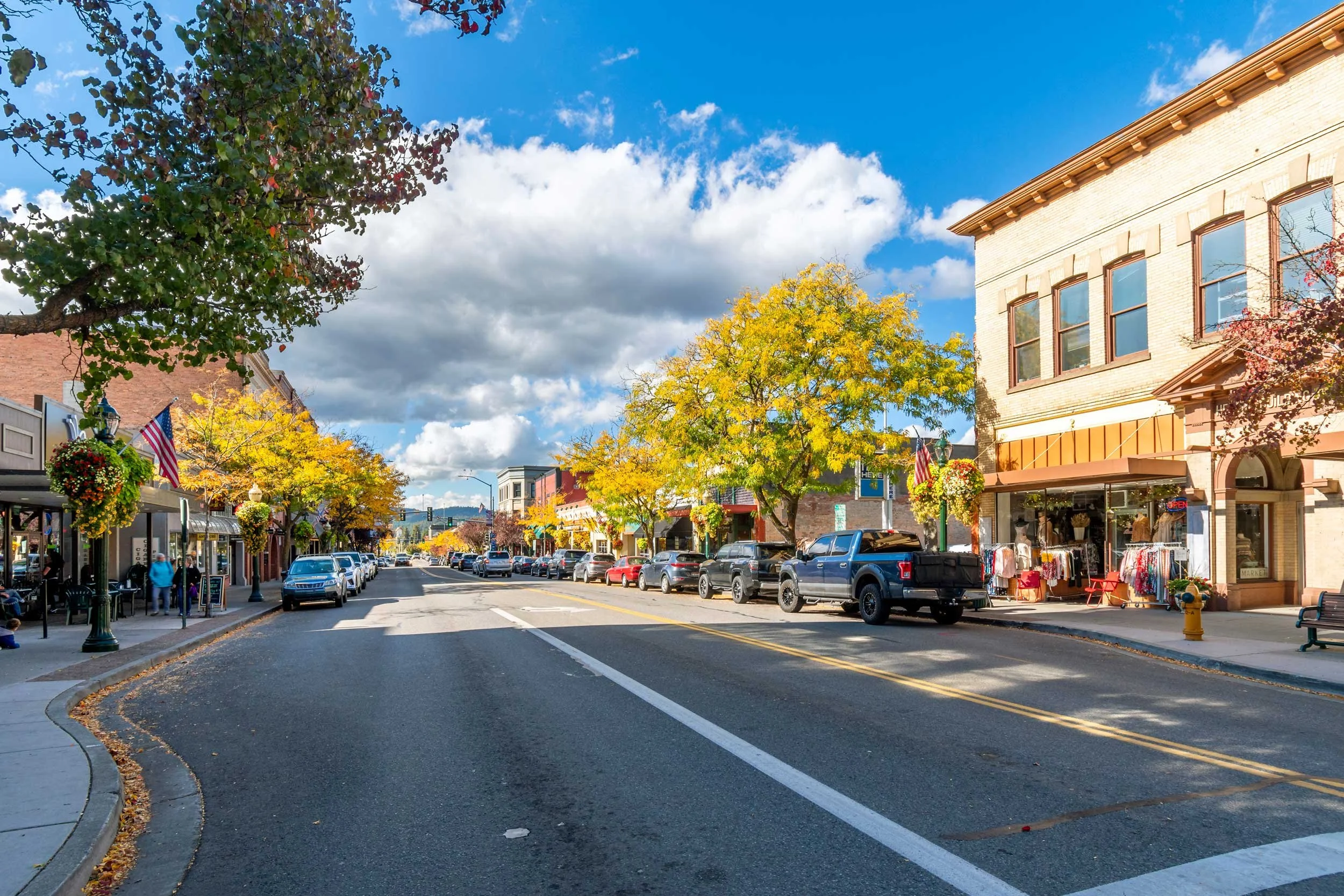 A small town street lined with colorful buildings and trees with autumn leaves, parked cars, and a clear blue sky with clouds.