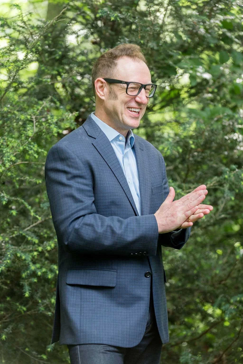A man in a suit smiling outdoors with greenery in the background.