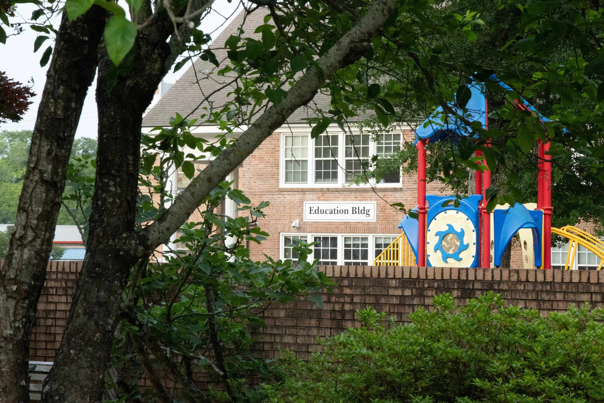 View of a brick school building with a sign reading 'Education Bldg,' surrounded by greenery and trees, with a colorful playground set partially visible on the right.