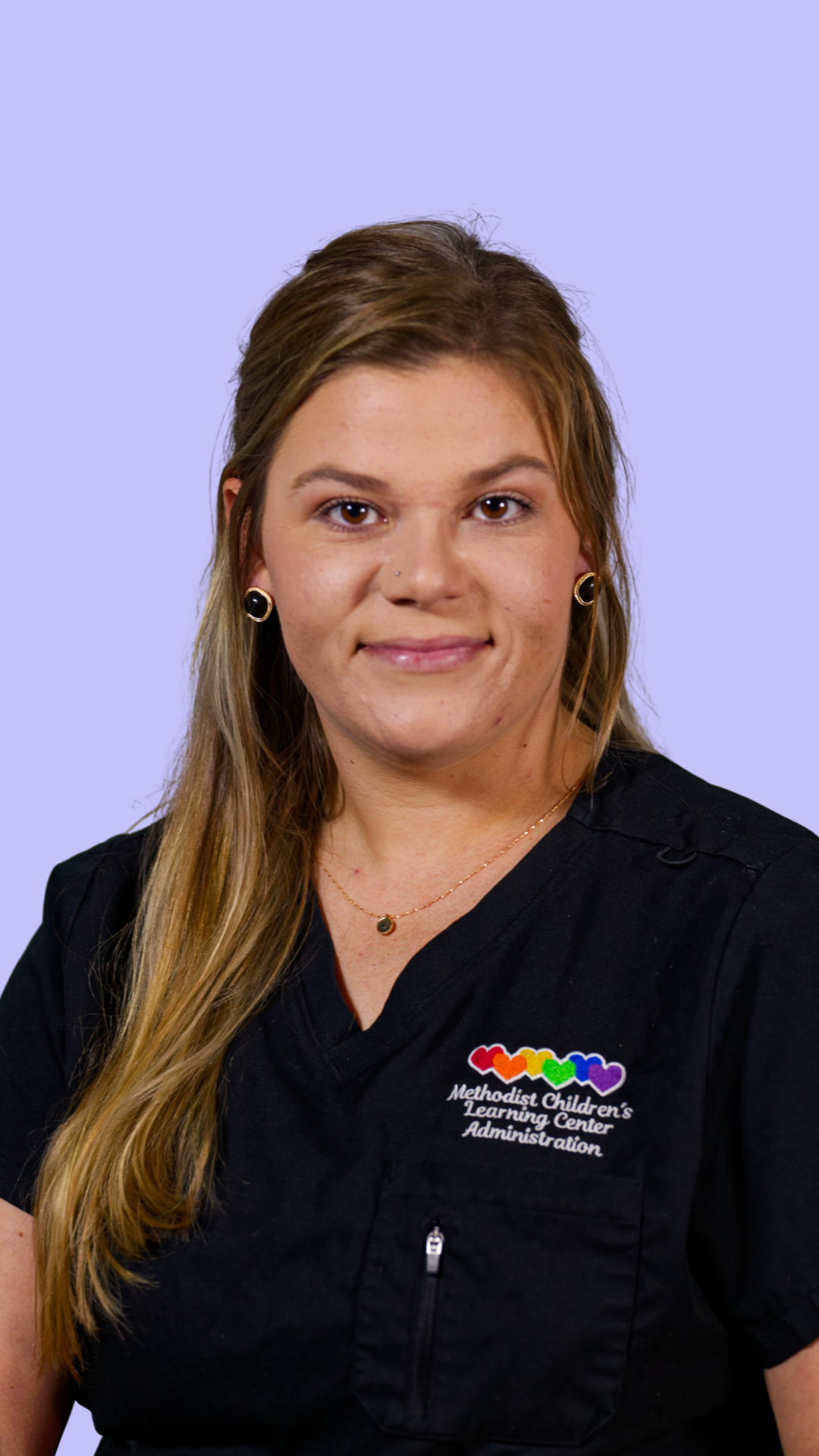 Portrait of a woman with long light brown hair, wearing a black scrub top with a colorful rainbow heart logo and the text 'Methodist Children's Learning Center Administration', gold earrings, and a delicate gold necklace, against a light purple background.