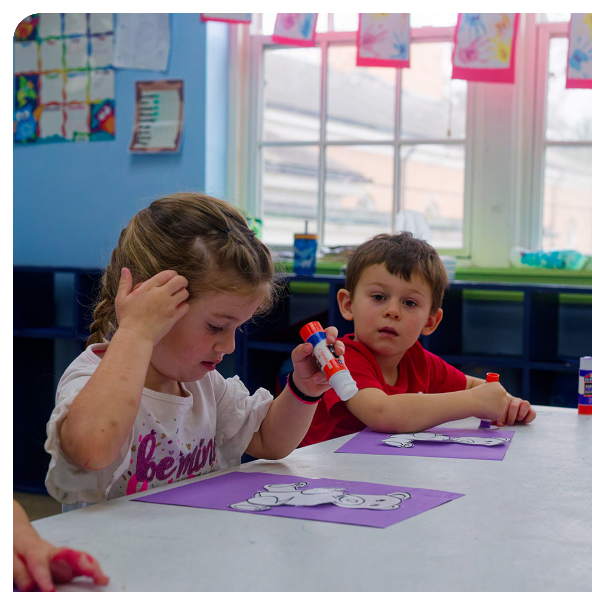 Children are sitting at a table engaged in arts and crafts, with purple papers and cut-out images of cartoon characters in front of them. The background shows a bright classroom with windows and colorful artwork on the walls.