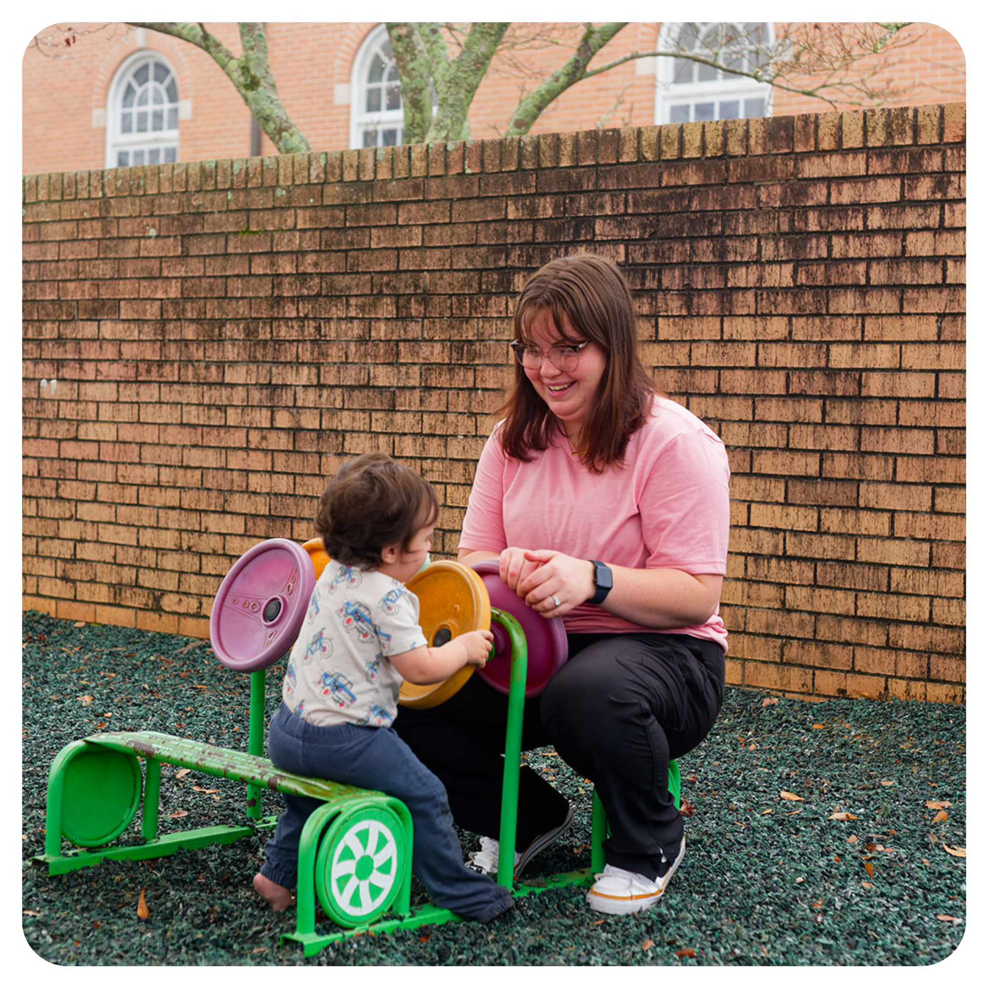A woman and a young child playing on a small colorful toy car outdoors, with a brick wall and a building with arched windows in the background.