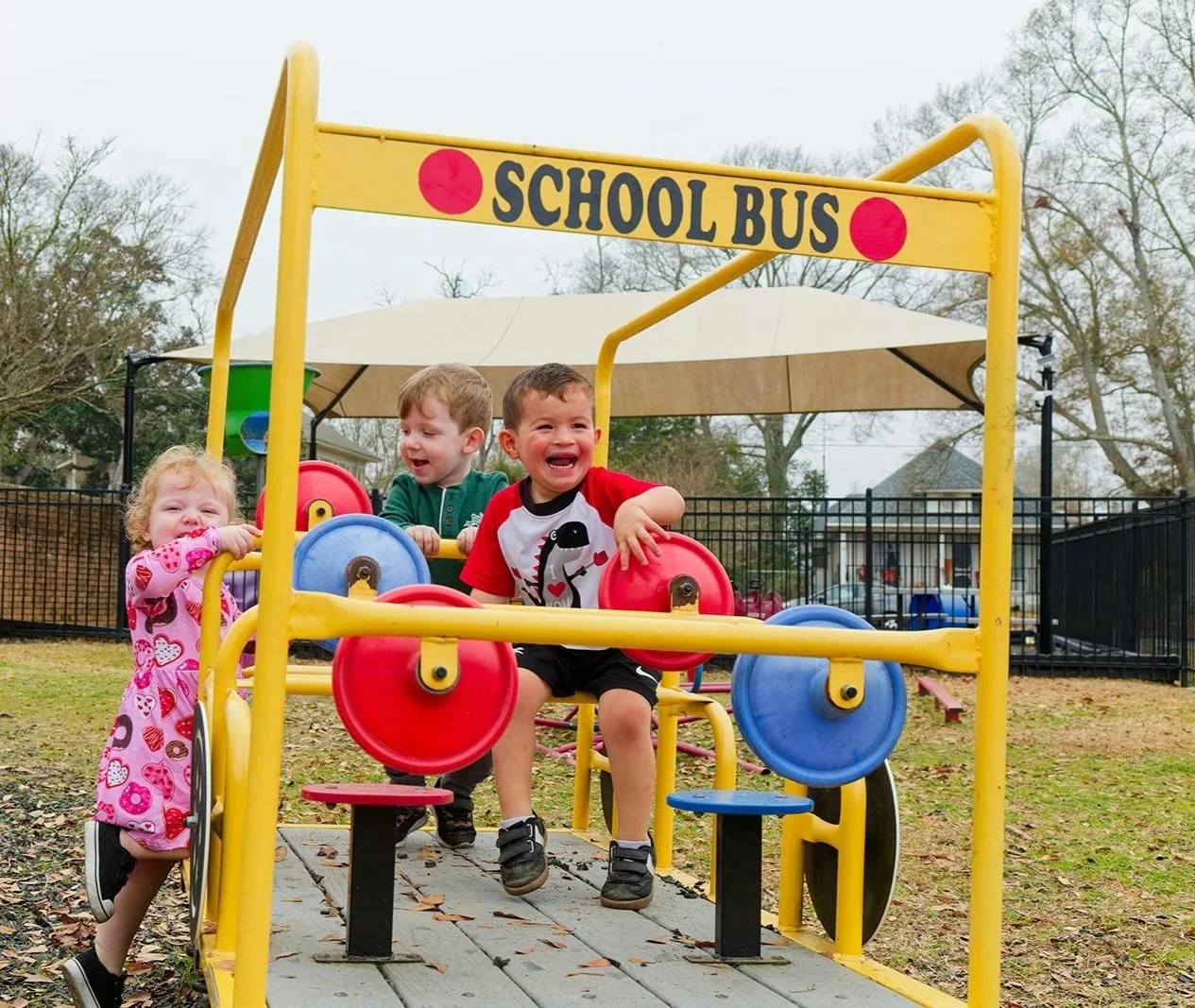 Children playing on a colorful outdoor playground with a yellow 'School Bus' structure, surrounded by leafless trees and a residential area.