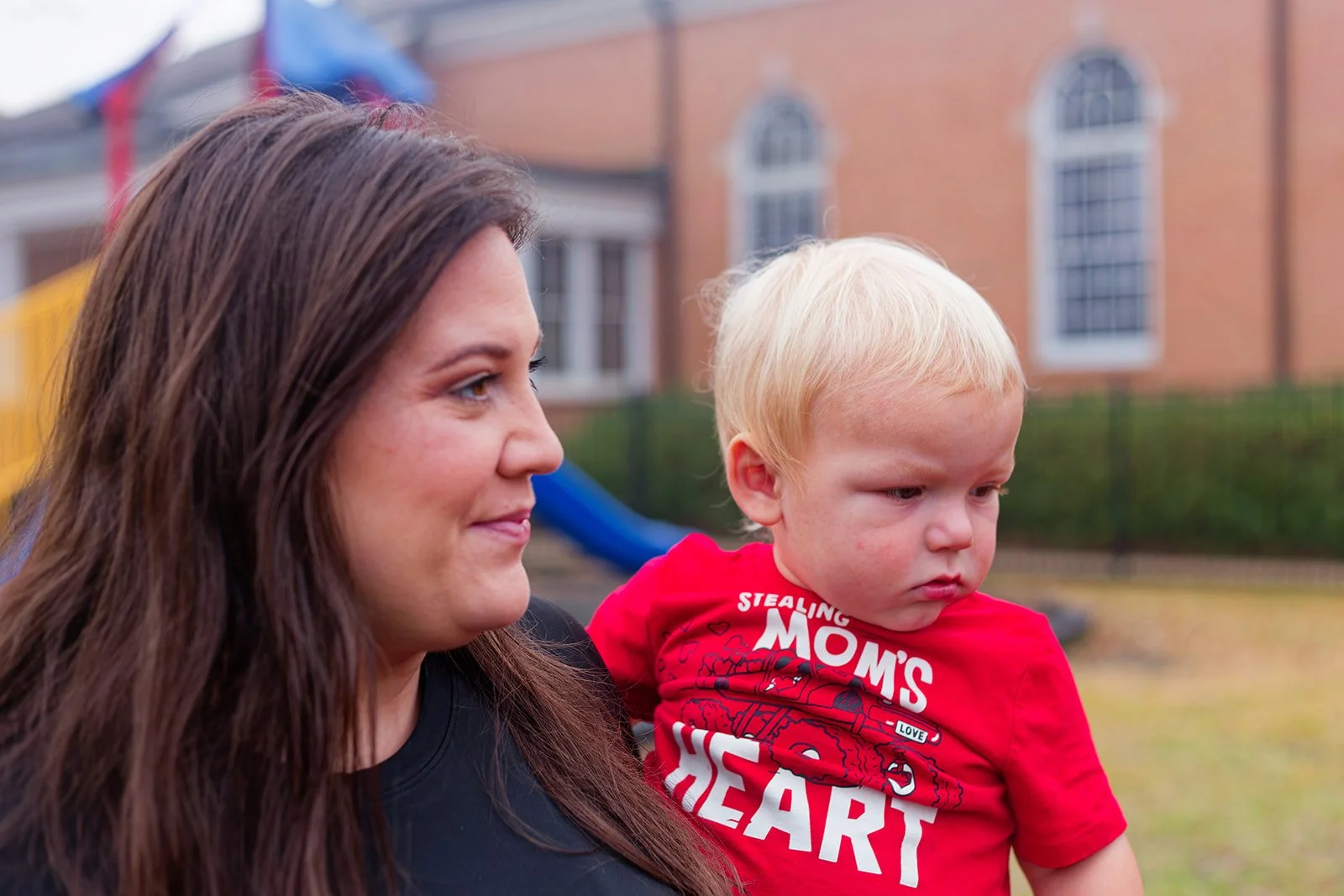 A woman with long dark hair holding a young blonde boy outdoors with a building and playground in the background.
