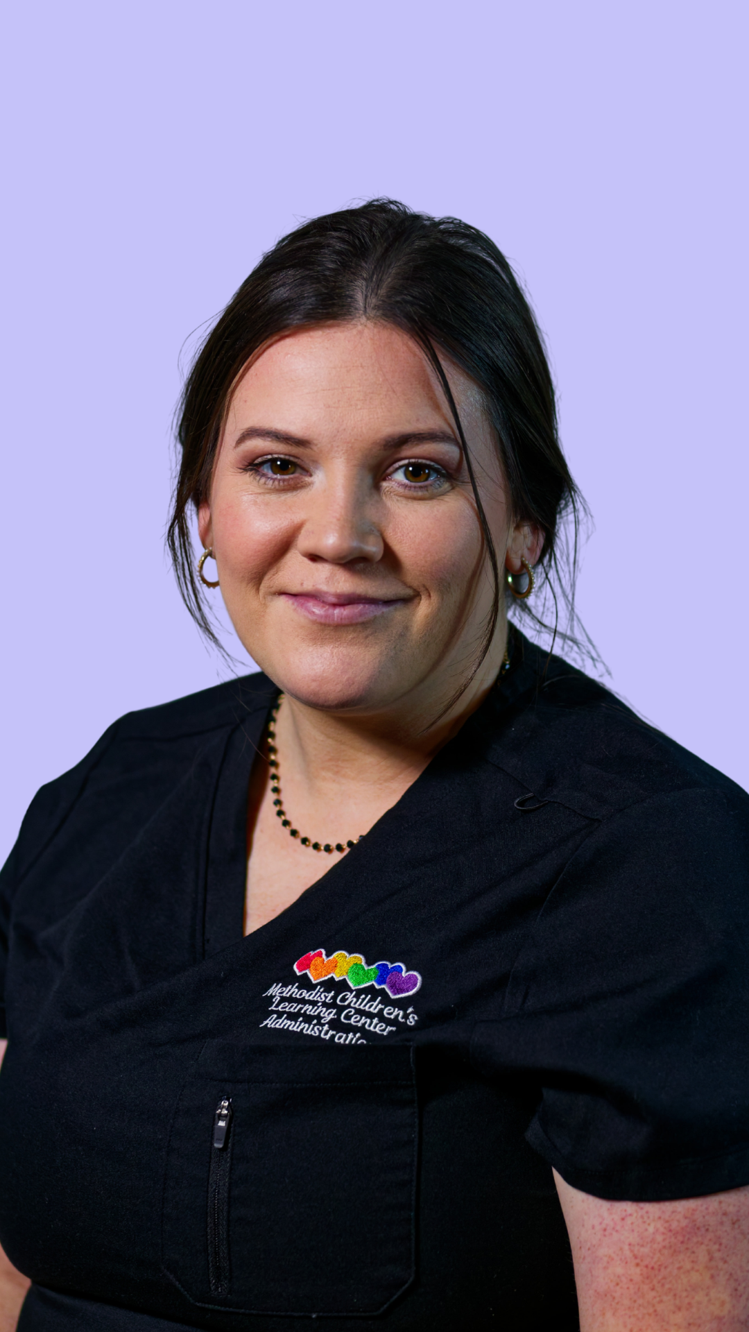 A woman with dark hair, wearing a black dress with a zipper and rainbow-colored logo for Methodist Children's Learning Center, smiles at the camera against a light purple background.