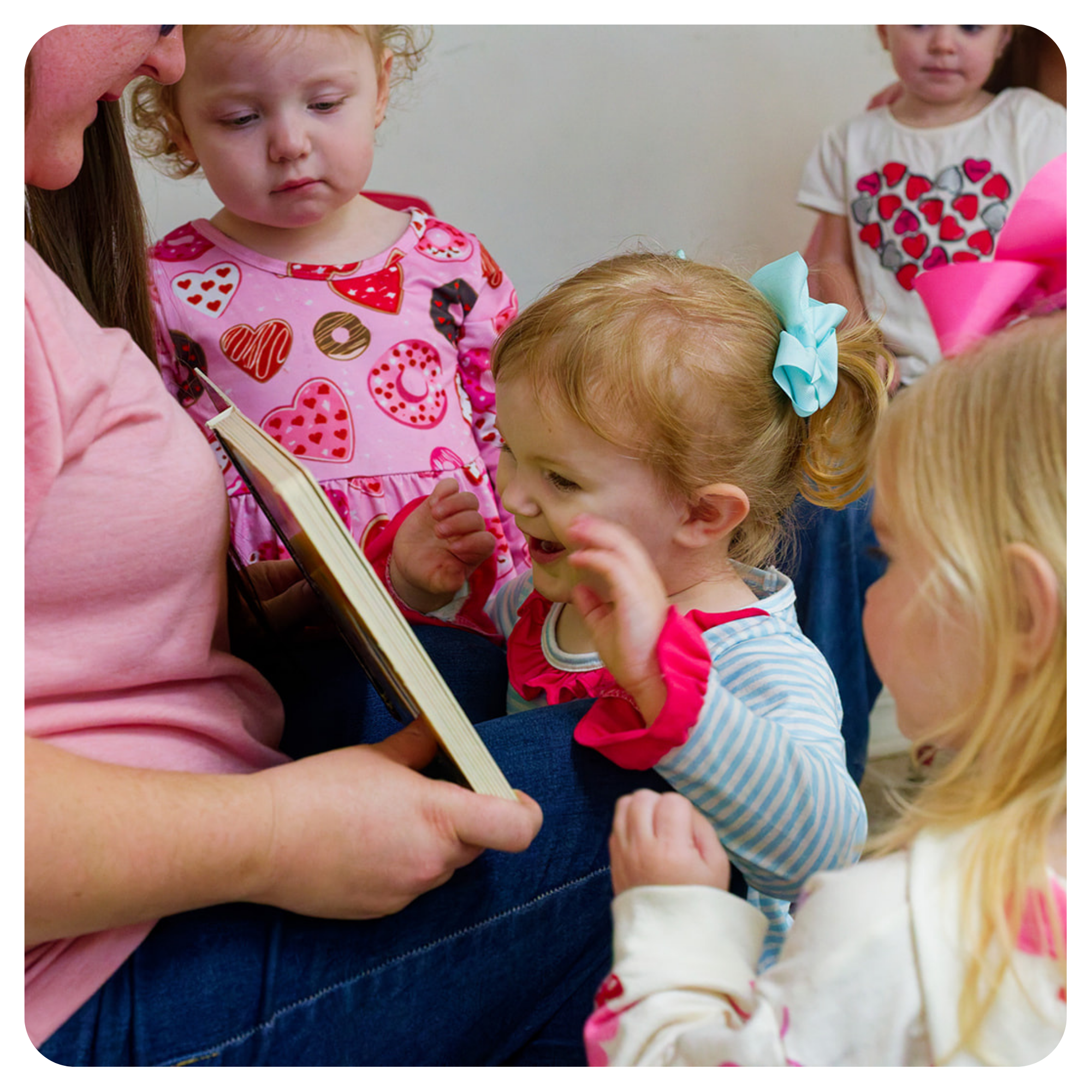 Group of young children gathered closely, some looking at a book held by an adult, with various facial expressions of curiosity and concentration.