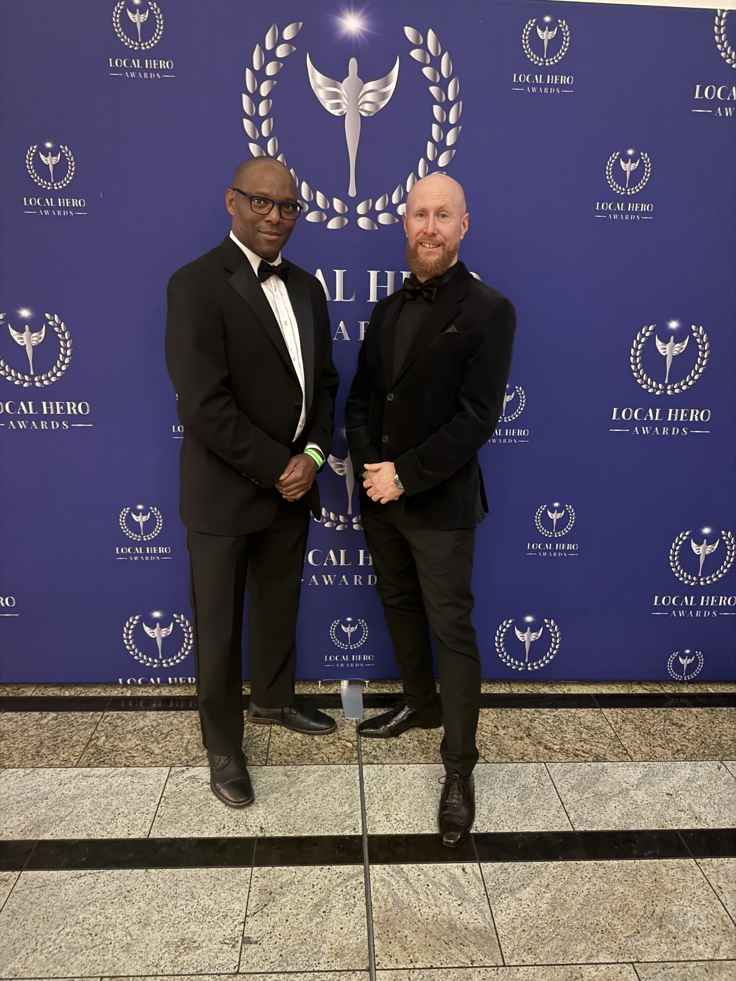 Two men in tuxedos standing in front of a blue backdrop with the 'Local Hero Awards' logo, at a formal event.