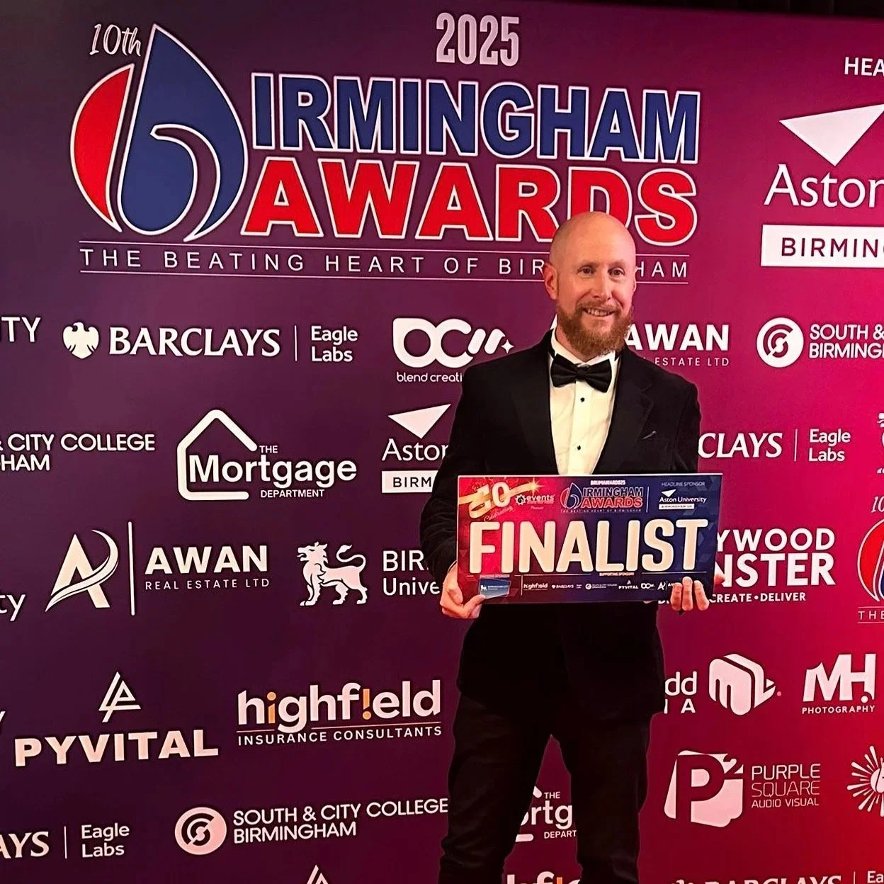 A man in a tuxedo holding a sign that says 'FINALIST' at the 2023 Birmingham Awards event, standing in front of a step-and-repeat backdrop with various sponsor logos.