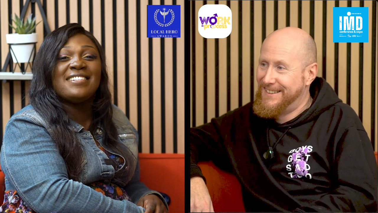 A woman with long dark hair and a denim jacket smiling, sitting next to a man with a beard and bald head wearing a black hoodie with text, in a setting with vertical wooden slats background and three logos at the top.