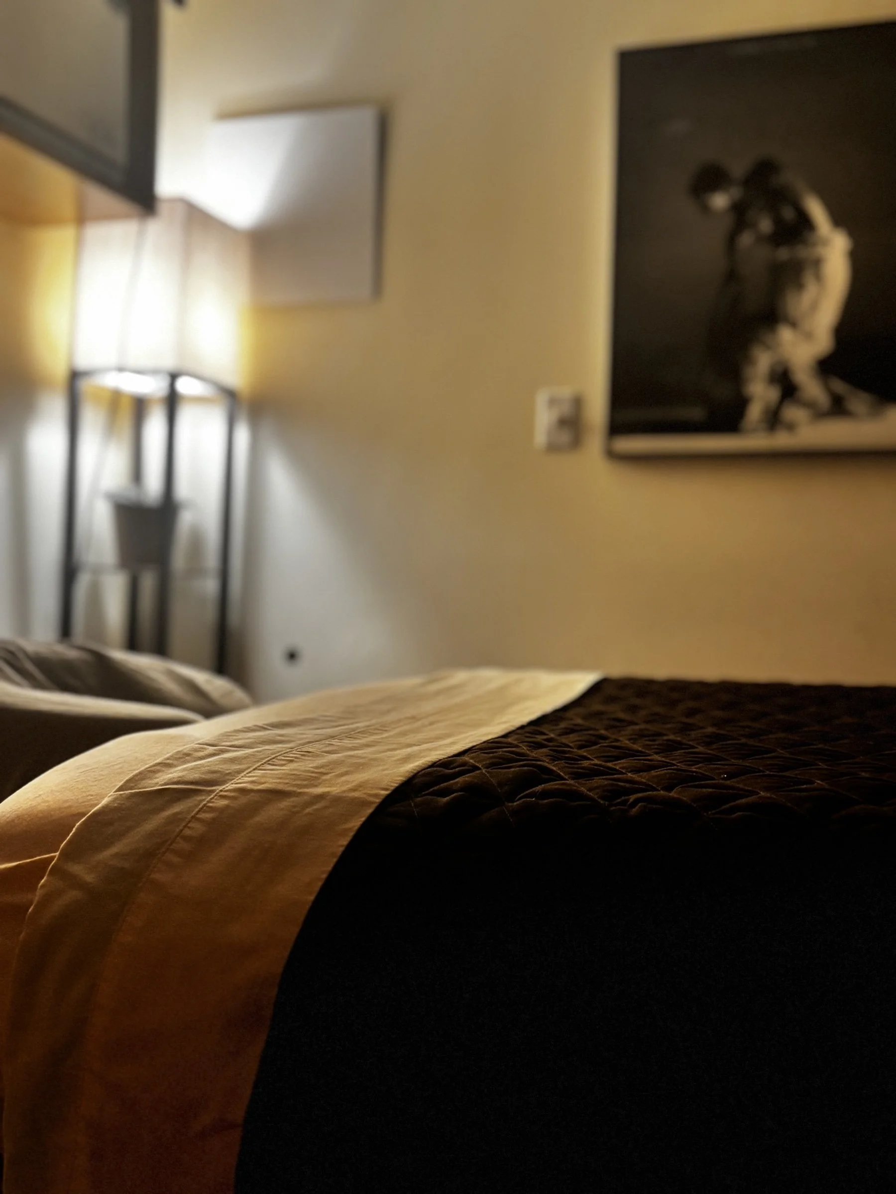 Close-up of a bed with a yellow and dark quilt, in a hotel room with a floor lamp and framed artwork on the wall.
