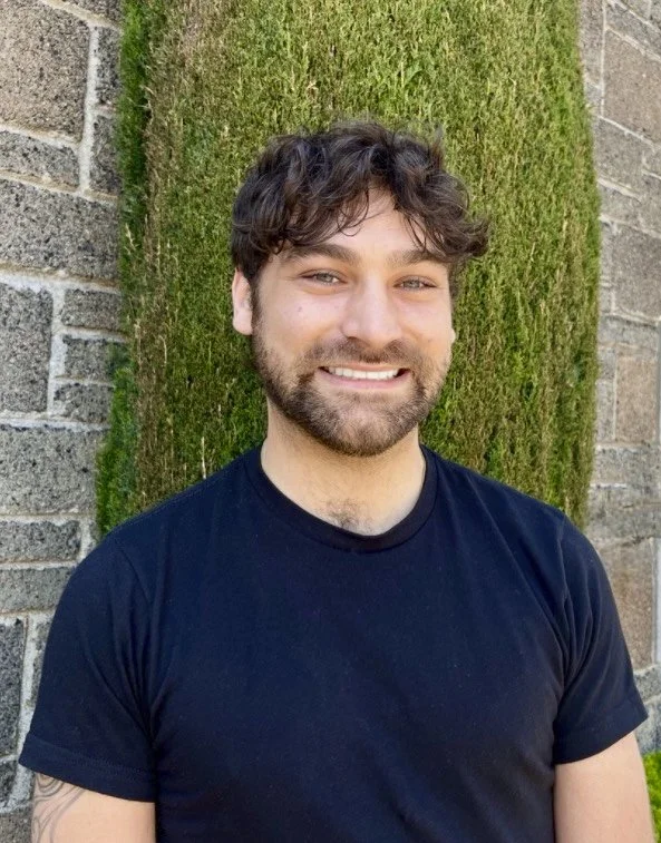 A smiling young man with curly dark hair and a beard, wearing a navy blue T-shirt, standing outdoors in front of a moss-covered brick wall.