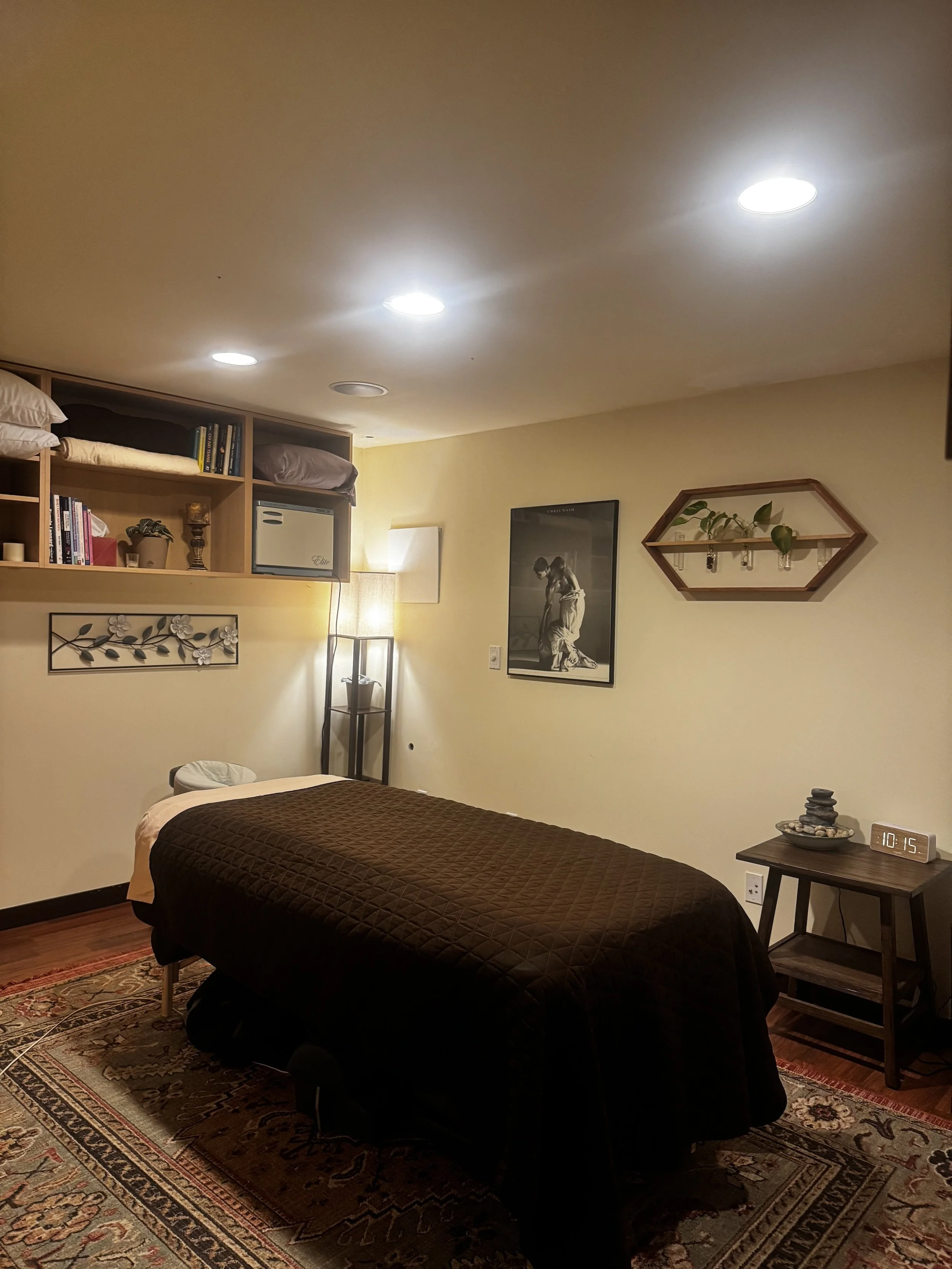 A bedroom with a bed covered in a dark bedspread, a wooden side table with a decorative bowl, a digital clock, and a lamp. The wall has framed artwork and a diamond-shaped shelf with a plant and hanging lights. There are books and pillows on a wall-mounted shelf and a colorful rug on the floor.