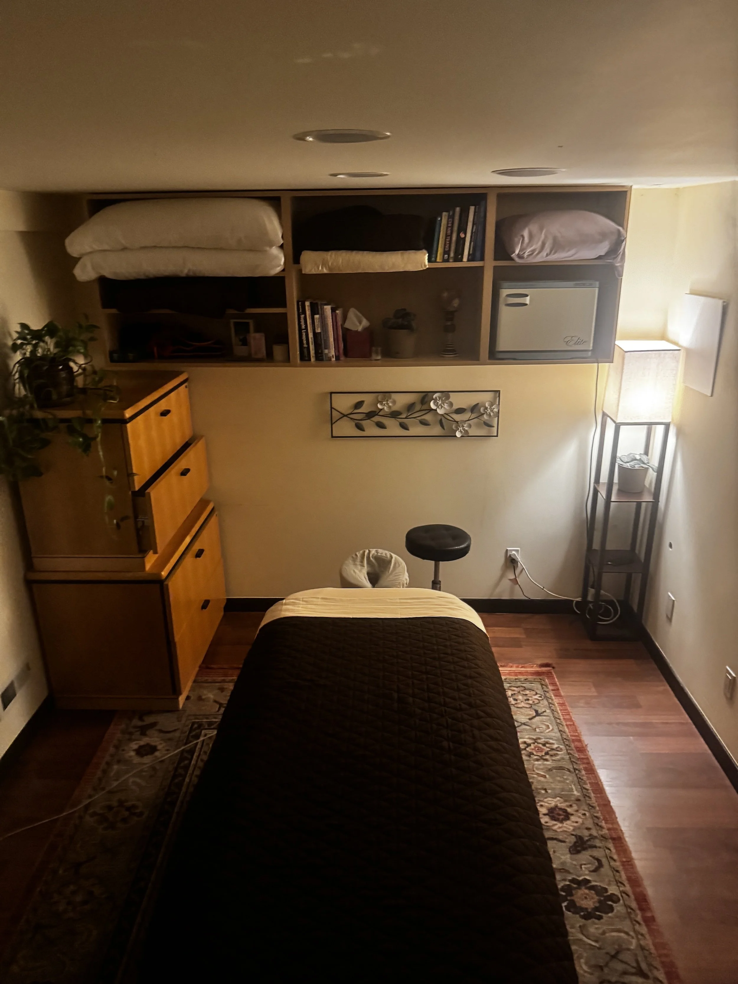 A bedroom with a bed in the center, a wooden dresser on the left, a bookshelf above the bed, a small safe on the right, a framed floral wall decor, a floor lamp on the right, a black stool, and a patterned area rug.