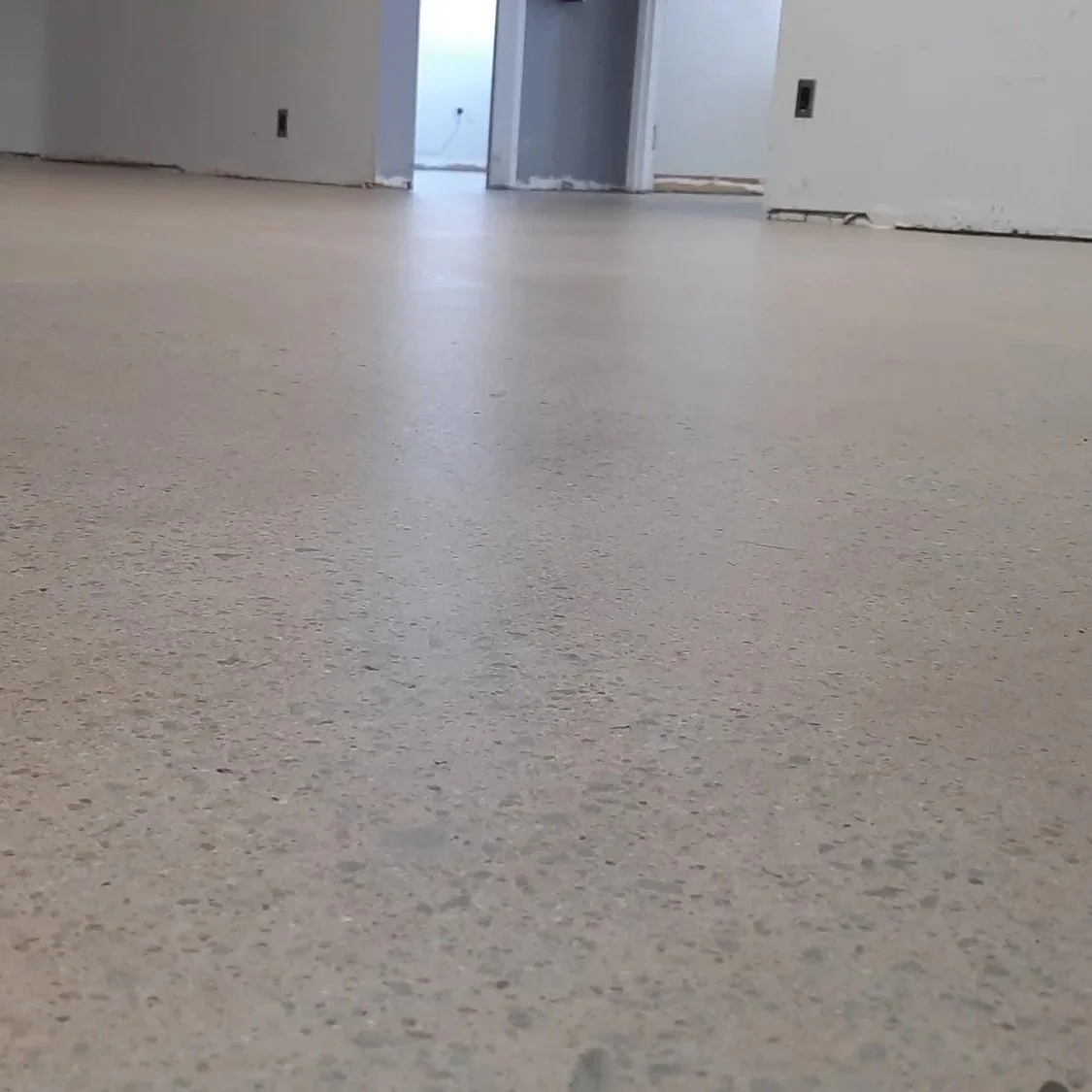 Interior view of a room with newly installed beige flooring, unfinished walls, and some electrical outlets. An open doorway reveals another room with natural light.