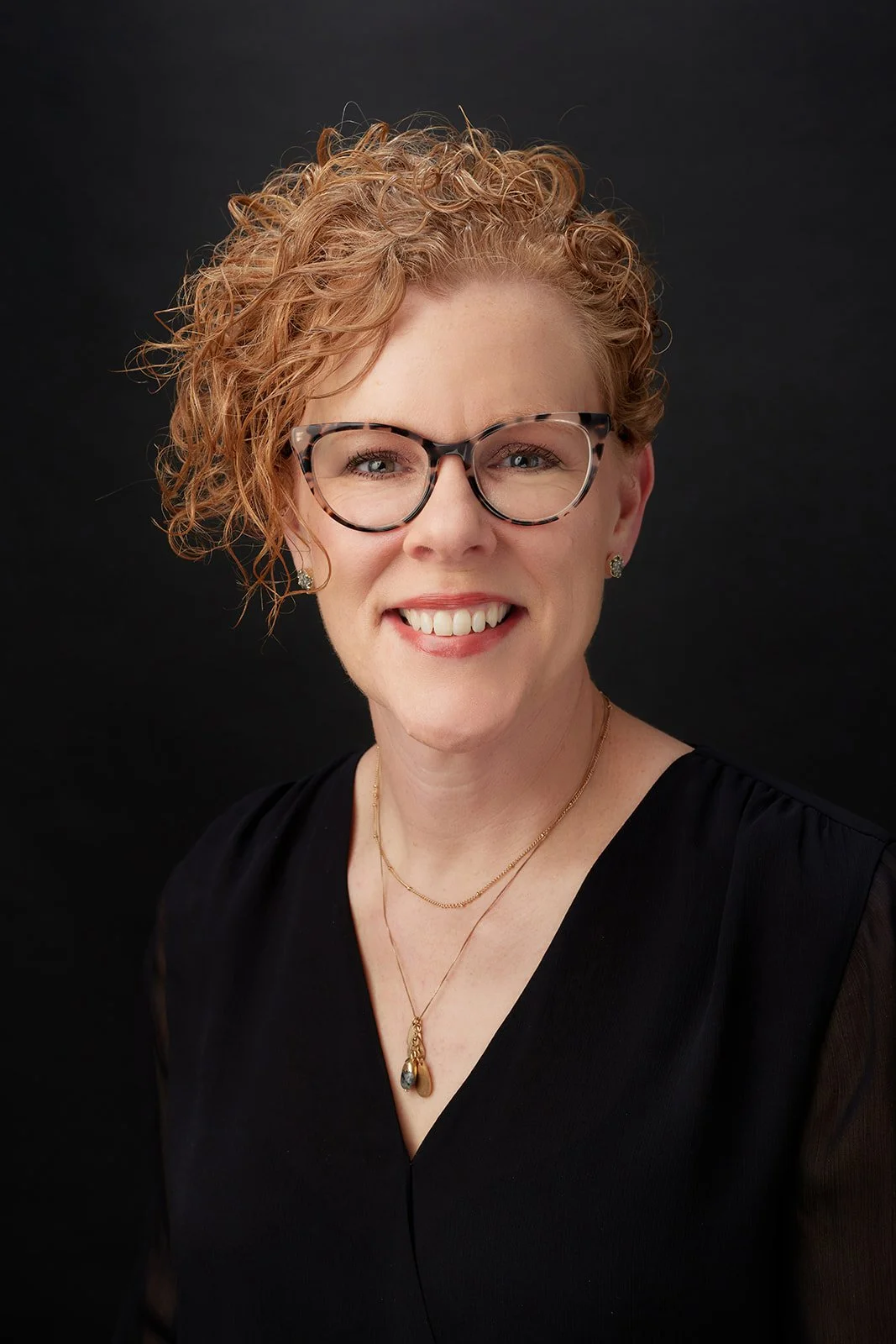 Portrait of a woman with short, curly red hair, wearing glasses, earrings, a black top, and layered gold necklaces, smiling against a black background.