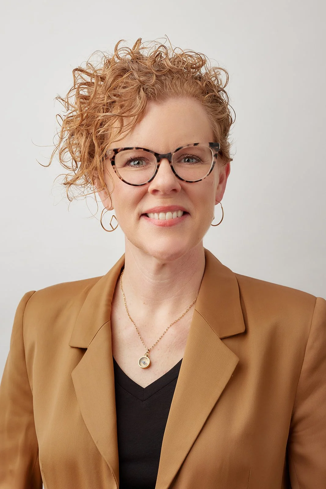 A woman with curly red hair, wearing glasses with tortoise shell frames, a tan blazer over a black top, and gold jewelry, smiling at the camera against a plain white background.