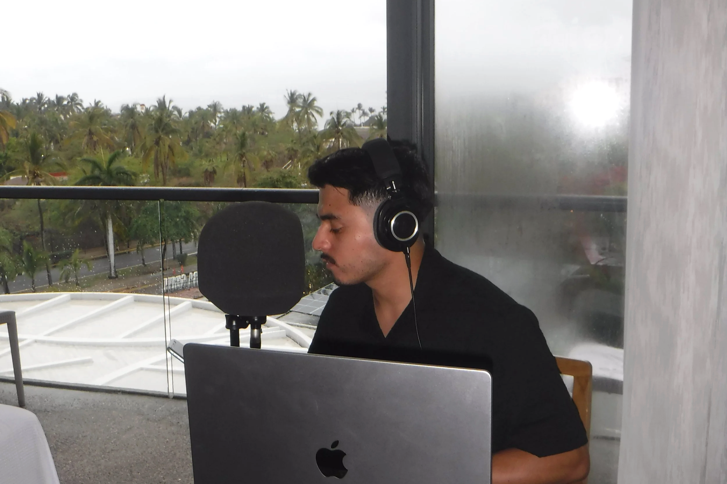 Aaron Tapia con auriculares grabando en un micrófono frente a una computadora portátil, con vista a palmeras y cielo lluvioso por una ventana.