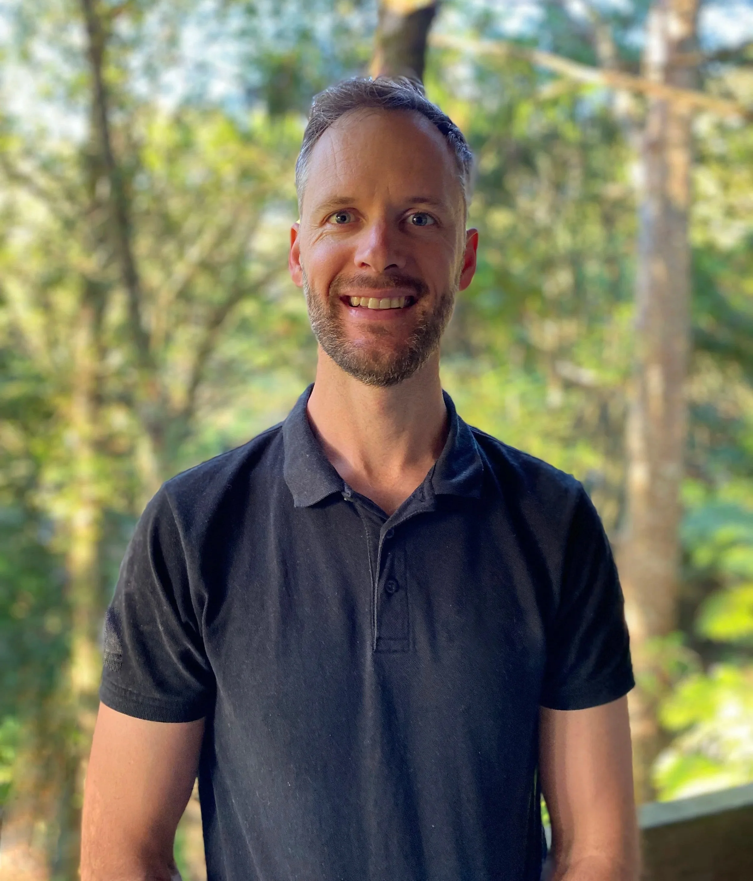 Close-up of a smiling man with blue eyes and a short beard, wearing a black shirt, outdoors with blurred green trees in the background.