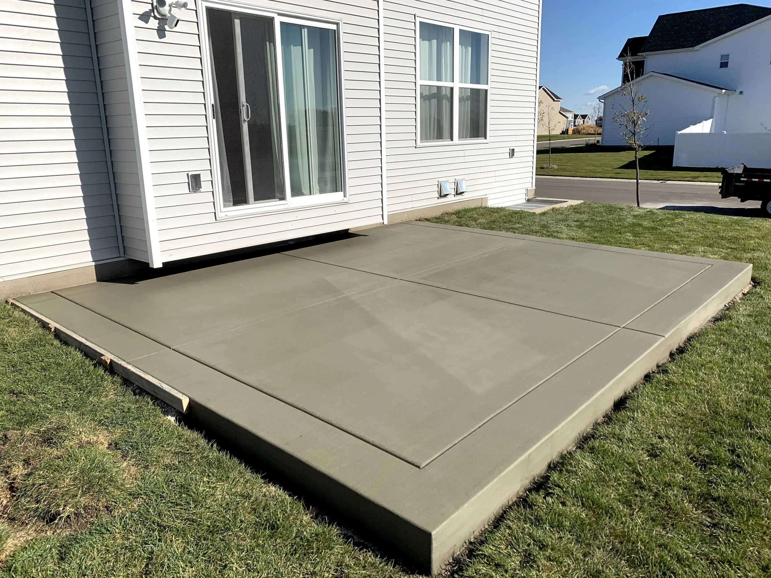 Newly poured concrete patio with defined edges and expansion joints, attached to the back of a house with white vinyl siding and sliding glass doors, surrounded by a green lawn in a suburban neighborhood.