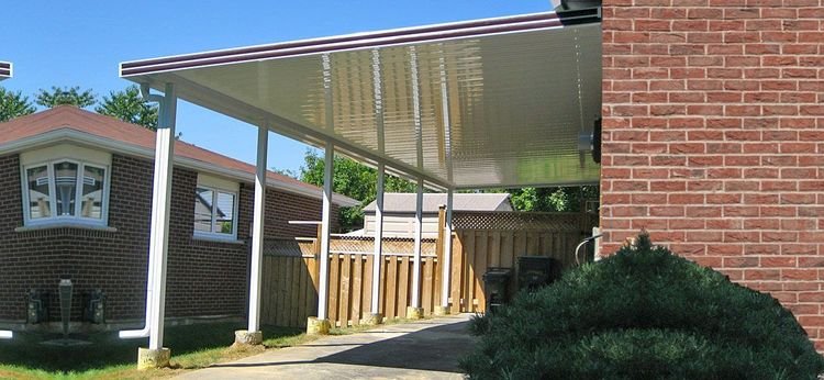 A covered outdoor patio area with a metal roof supported by white metal posts, adjacent to a brick house, with a wooden fence and some bushes in the background.