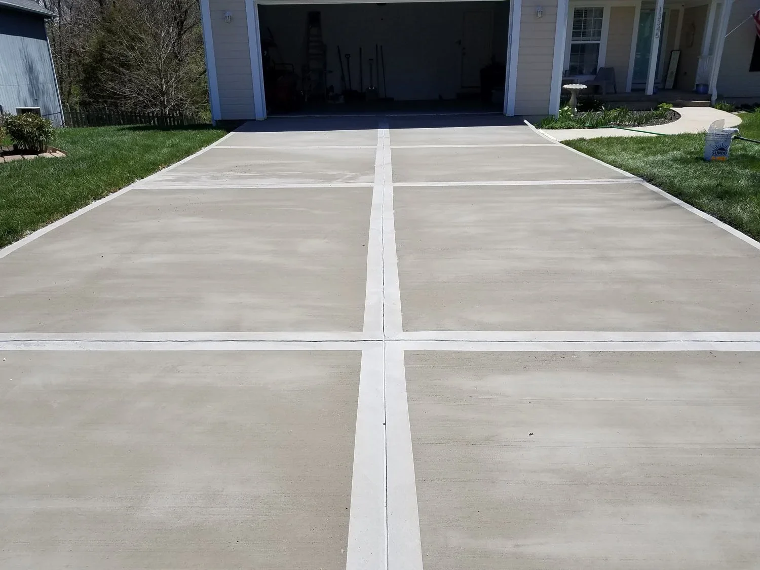 Empty concrete driveway with white borders leading to garage, flanked by grass and a house in the background.