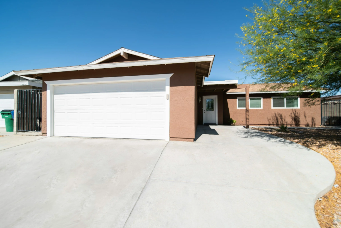 Front exterior of a single-story house with a white garage door, a beige stucco facade, and a small porch area. There is a large tree with green leaves on the right and a clear blue sky overhead.