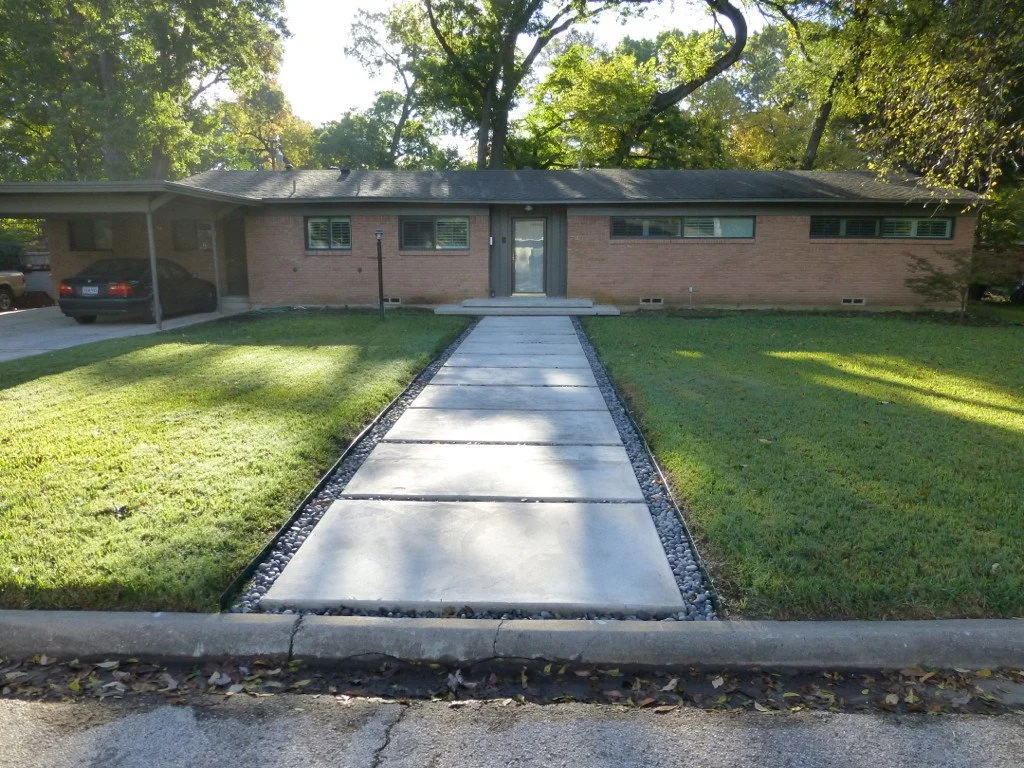 Concrete walkway leading to a brick house with a sidewalk in front. Lawn on both sides, trees in the background, and a car parked under a carport.
