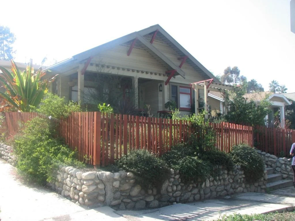 This double gabled Craftsman at 4866 Lincoln Ave is surrounded by a beautiful arroyo stone wall that is topped by a simple wooden picket fence. The avocado green clapboard siding has a horizontal one over two pattern. A darker green and crimson trim 