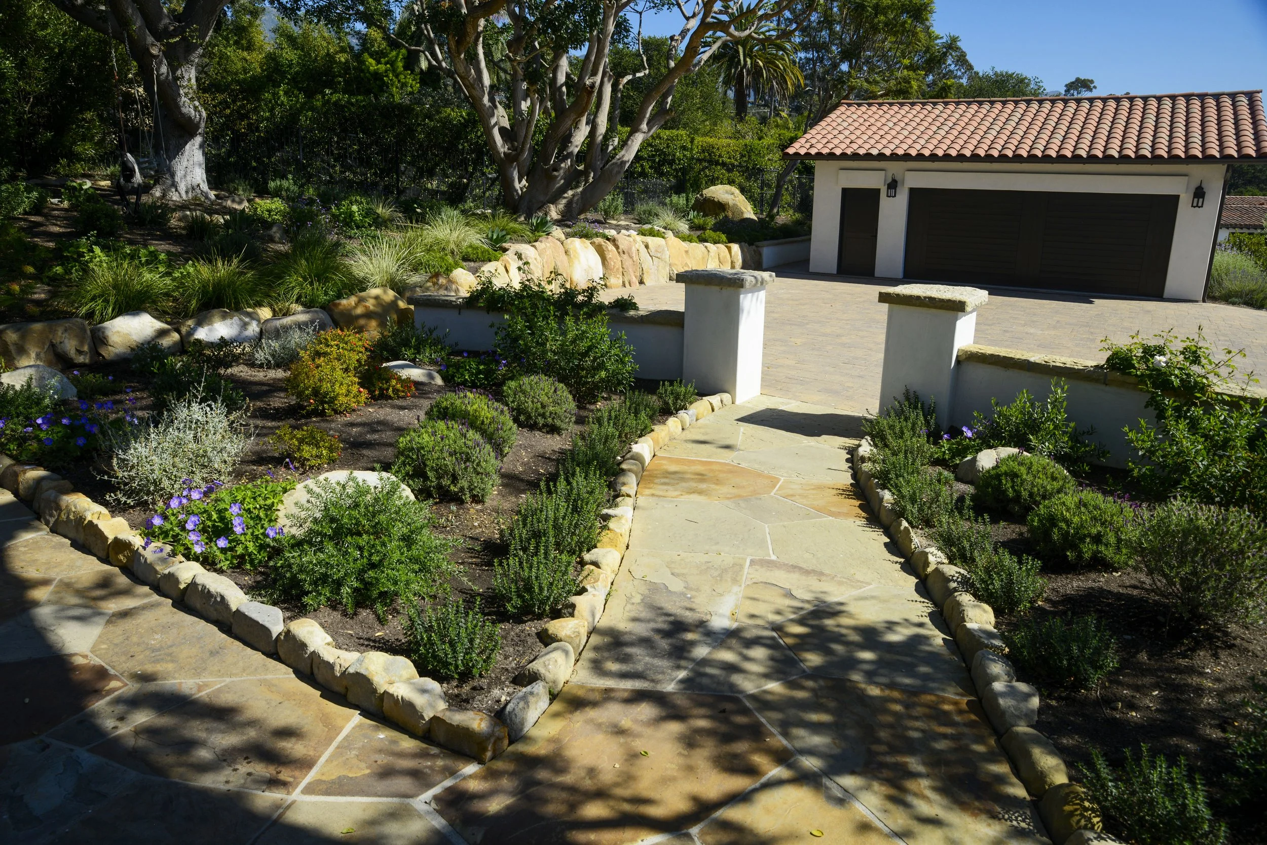 A landscaped front yard with a stone pathway, bordered by plants and flowers, leading to a garage with a tiled roof in the background.