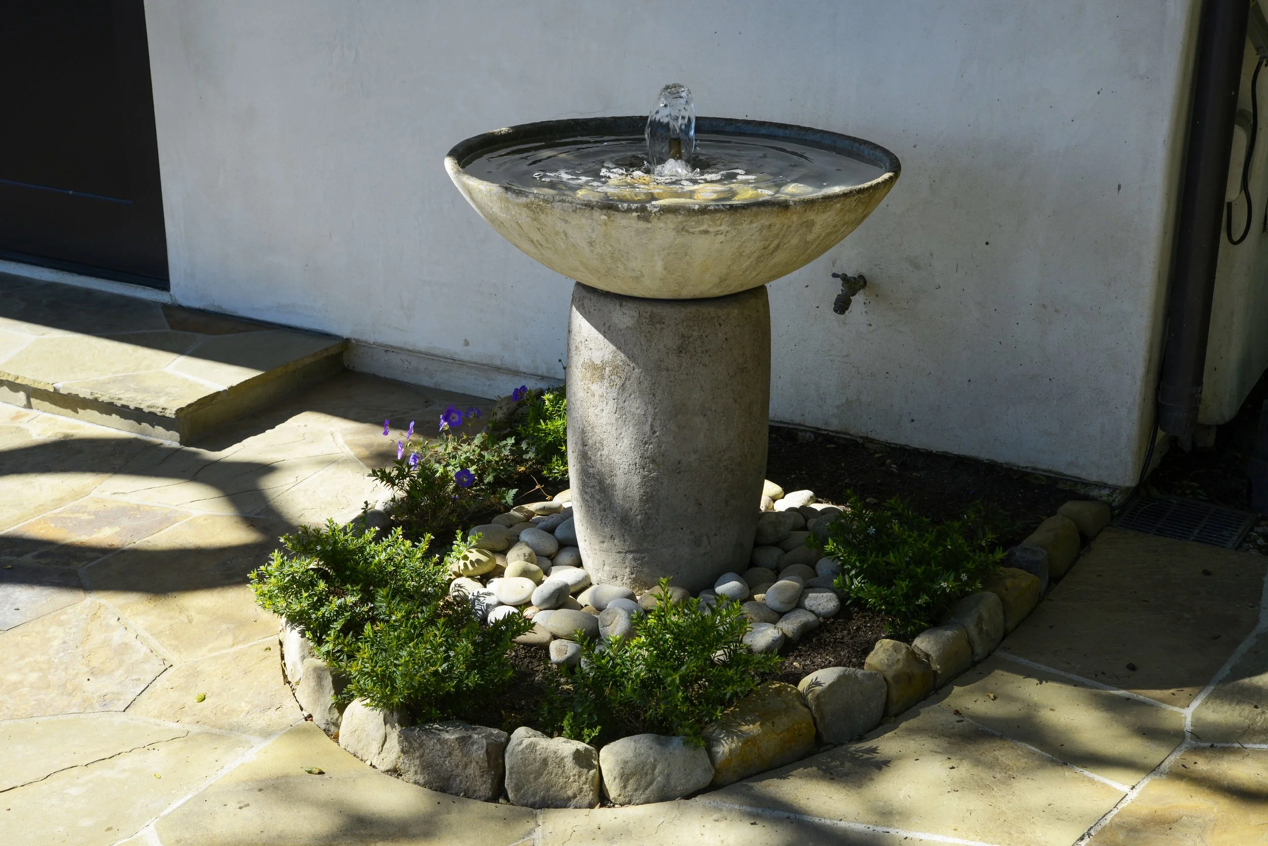 Outdoor garden birdbath with water spouting from a small fountain, surrounded by small plants, rocks, and paving stones.