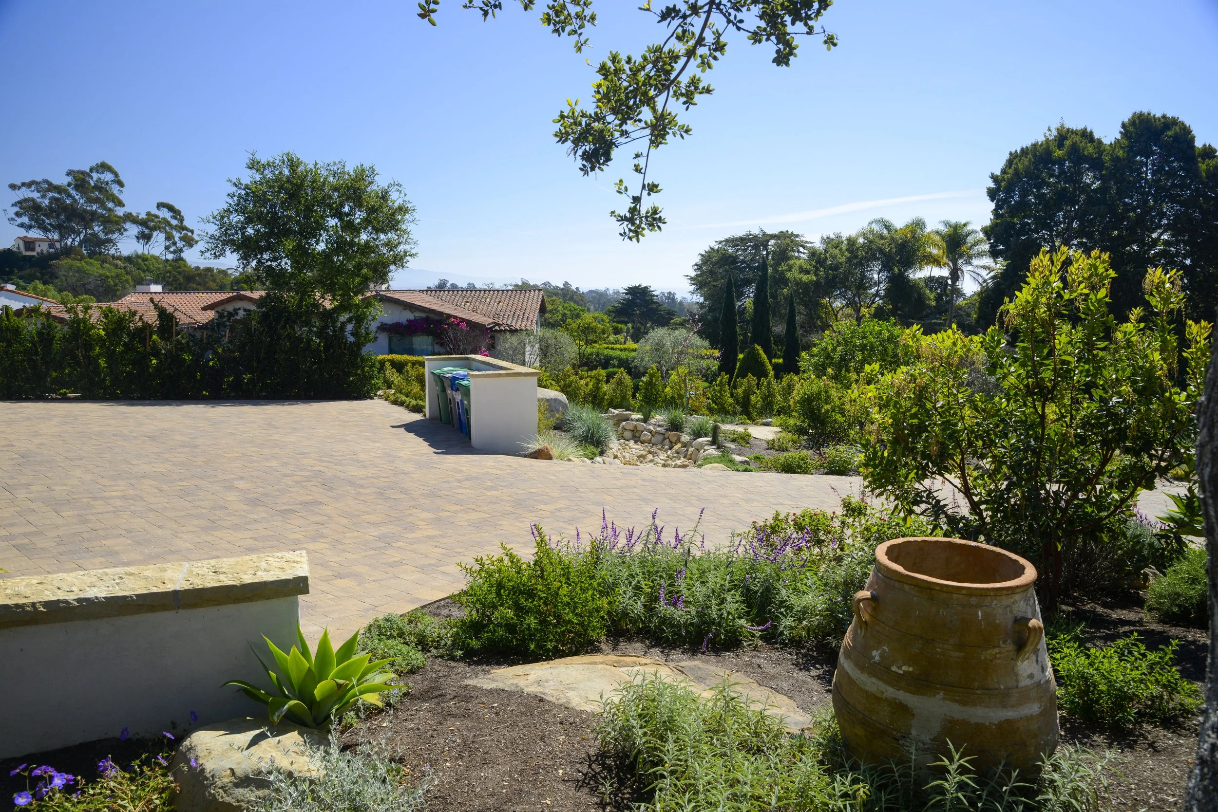 A spacious yard with succulent plants and gardens, a large empty clay pot, and a paved area with refuse bins, surrounded by lush green trees under a clear blue sky.