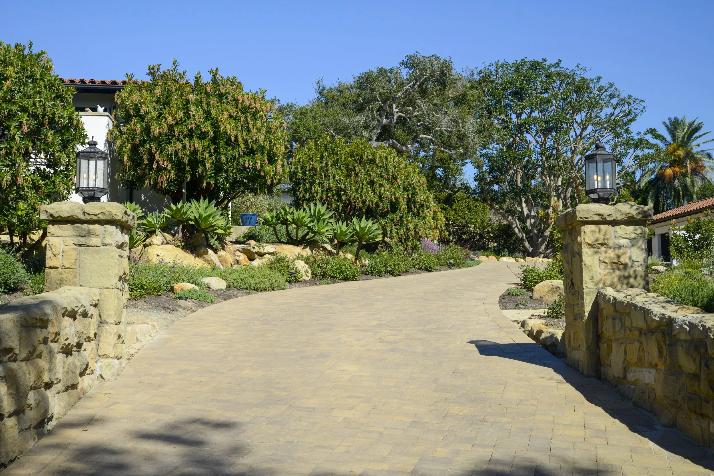 A curved stone pathway in a lush garden, flanked by stone pillars with lanterns, surrounded by various green trees and plants, under a clear blue sky.