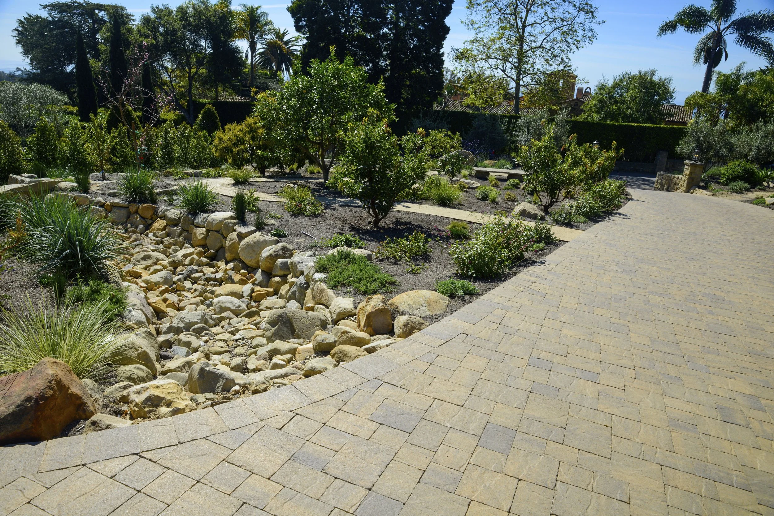 A landscaped garden with various plants and trees, featuring a winding stone pathway and a dry creek bed with rocks.