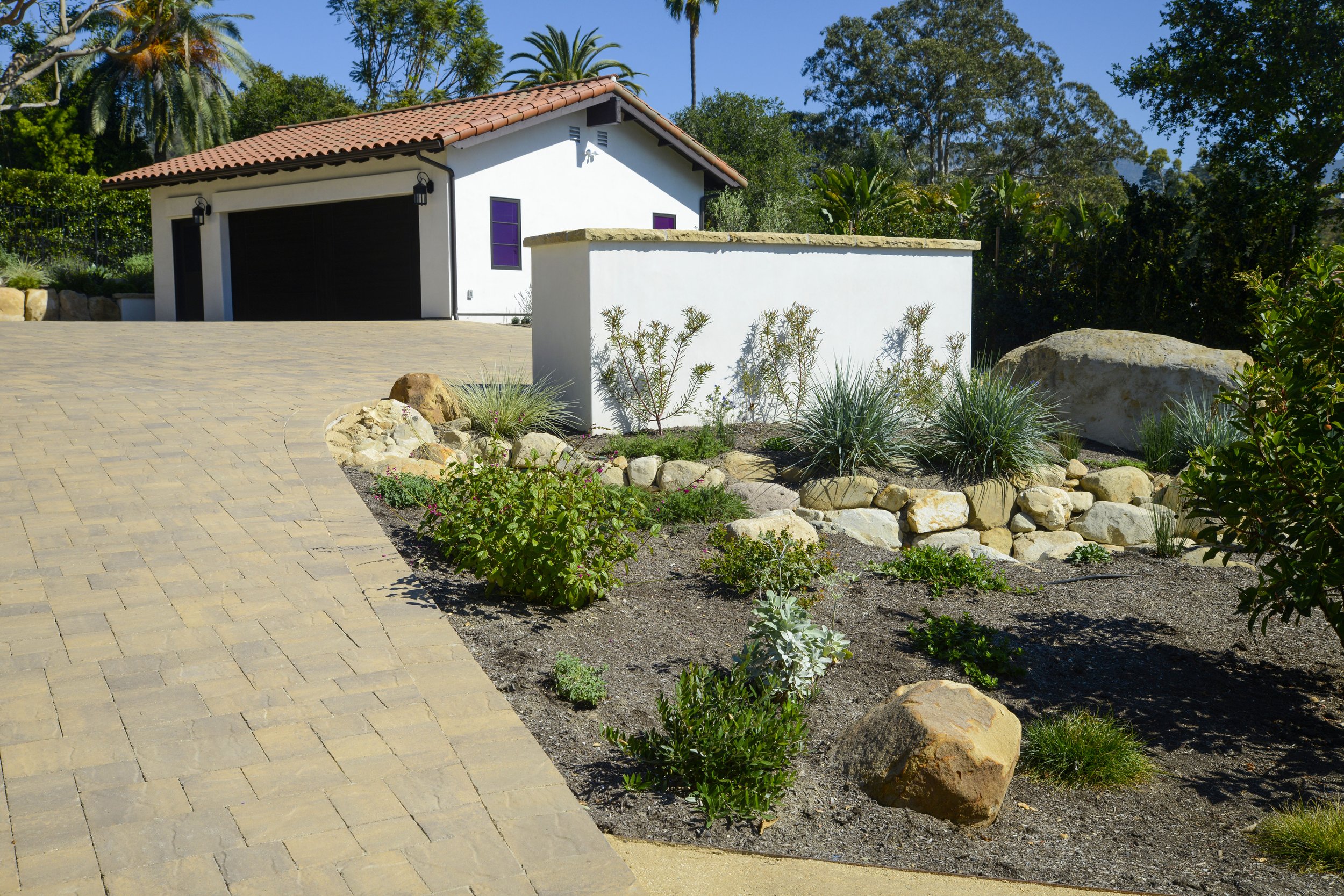 A beige stone-paved driveway leads up to a white house with a red tiled roof, surrounded by desert landscaping with rocks and drought-resistant plants, and lush green trees in the background under a clear blue sky.