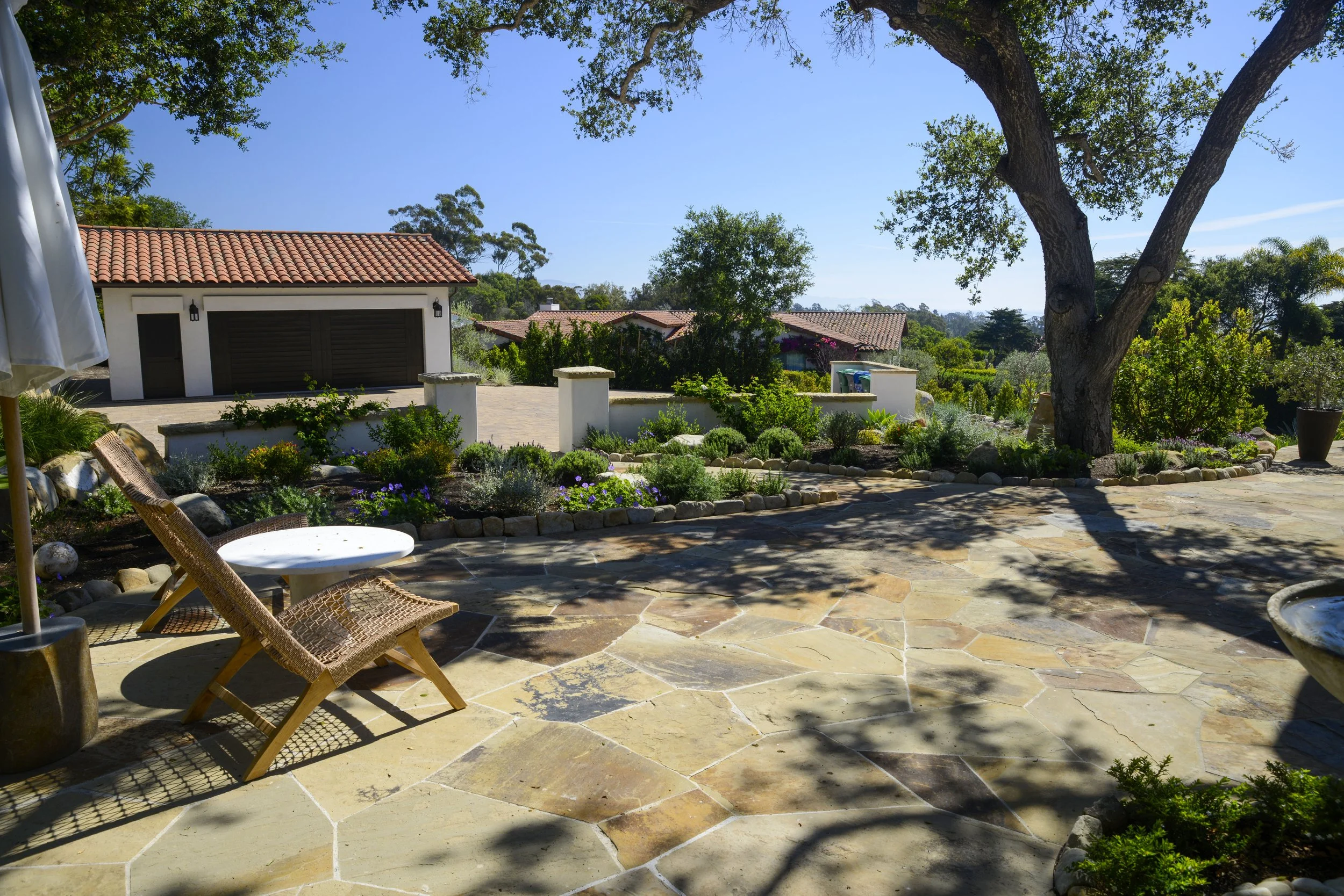 A sunny backyard patio with stone flooring, outdoor chairs, a white round table, a large shadow of a tree, a garden with plants and flowers, a large tree, and a house with a garage and red tile roof in the background.