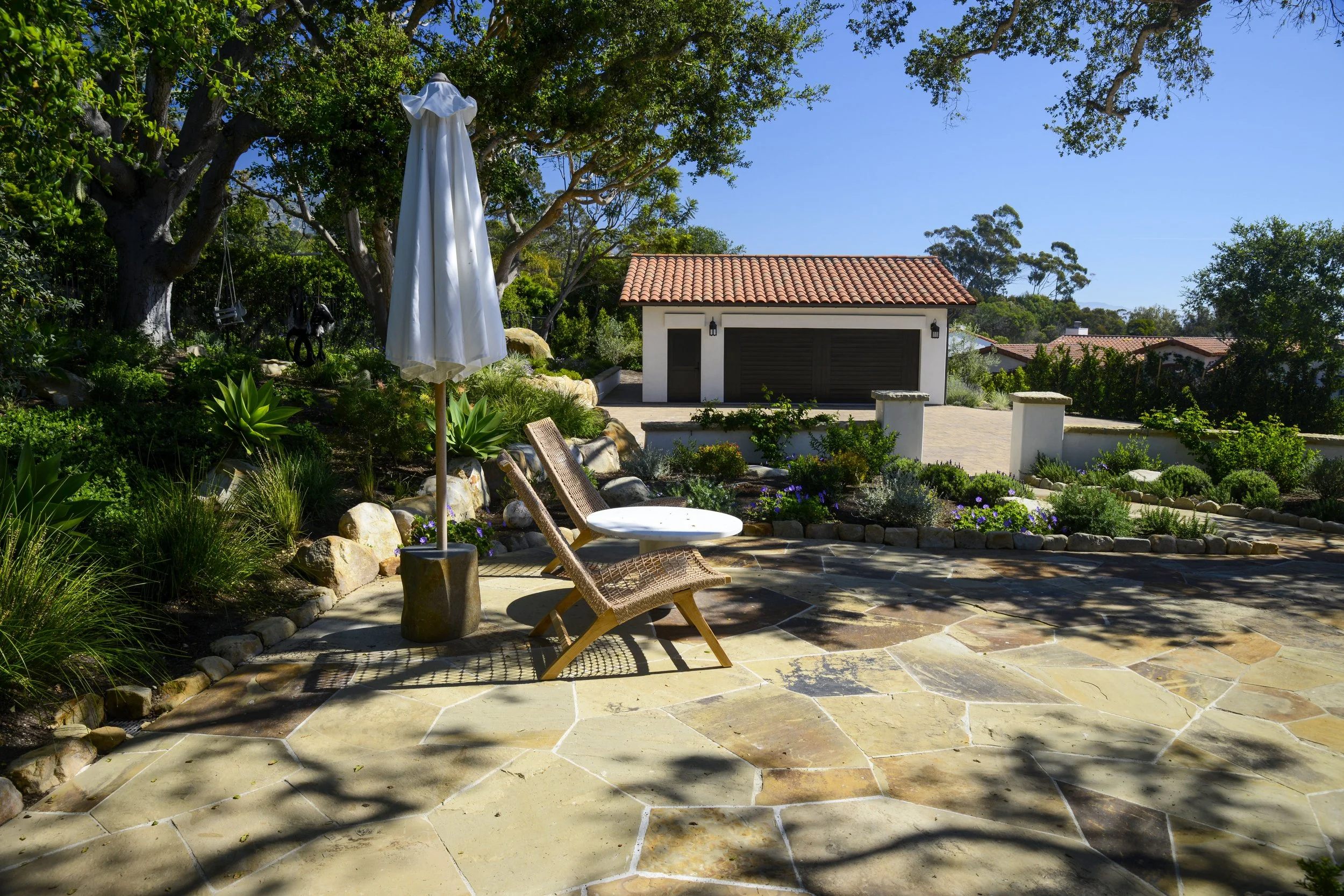 A backyard patio with two wicker chairs and a small round table under a closed white patio umbrella, surrounded by trees, plants, and a landscaped garden with flowers, rocks, and a distant garage with a red tiled roof.